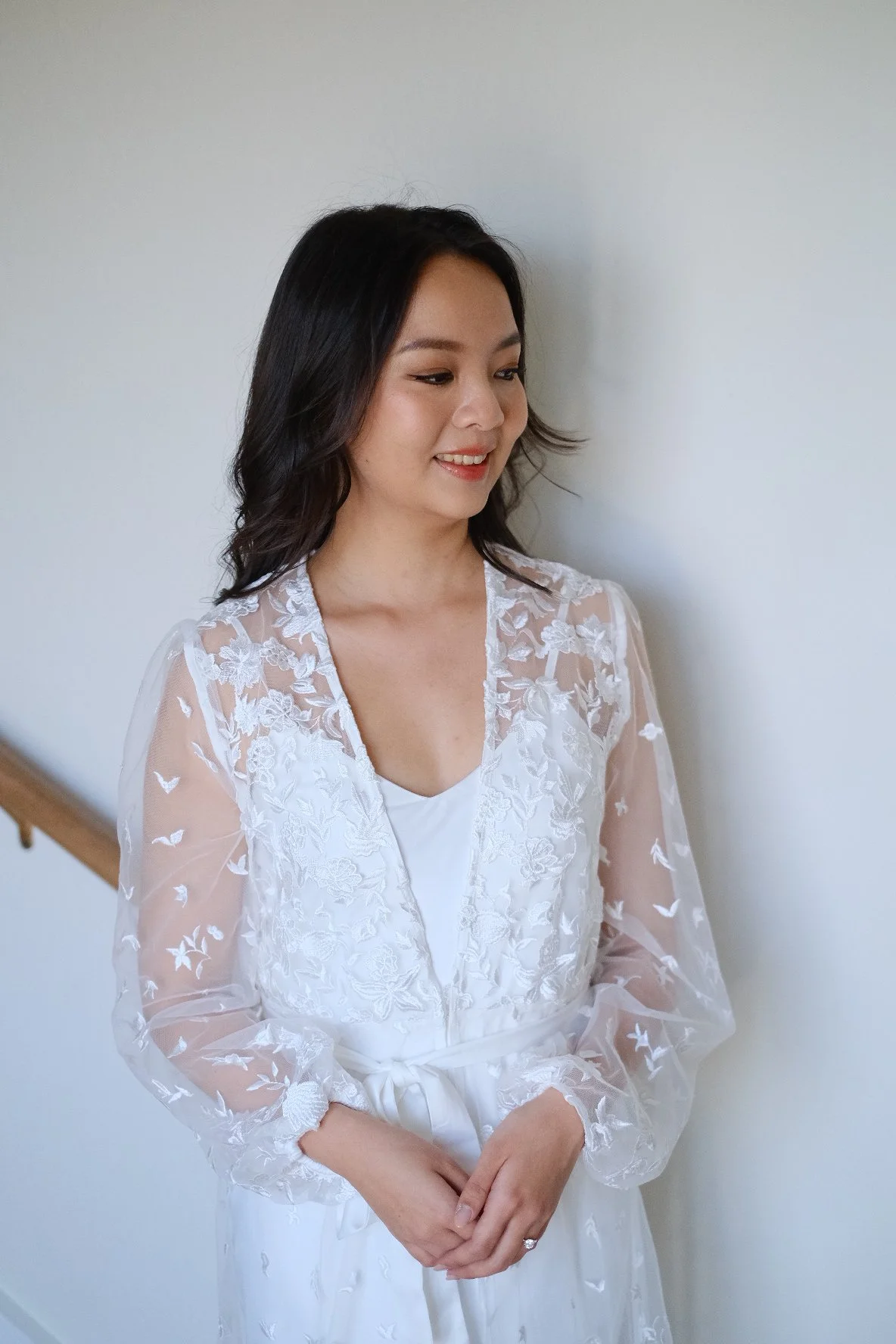 A woman with black hair wearing a white dress with sheer floral embroidery, standing against a plain wall.