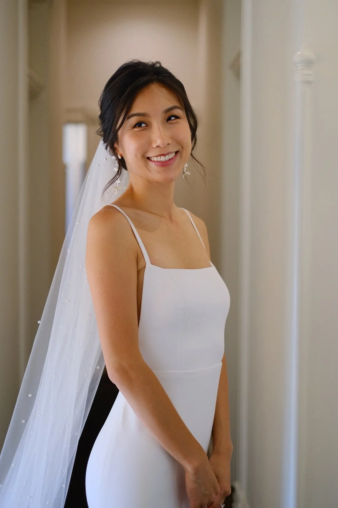 A woman in a white dress with spaghetti straps, smiling, wearing earrings and a veil, standing indoors near a corner.