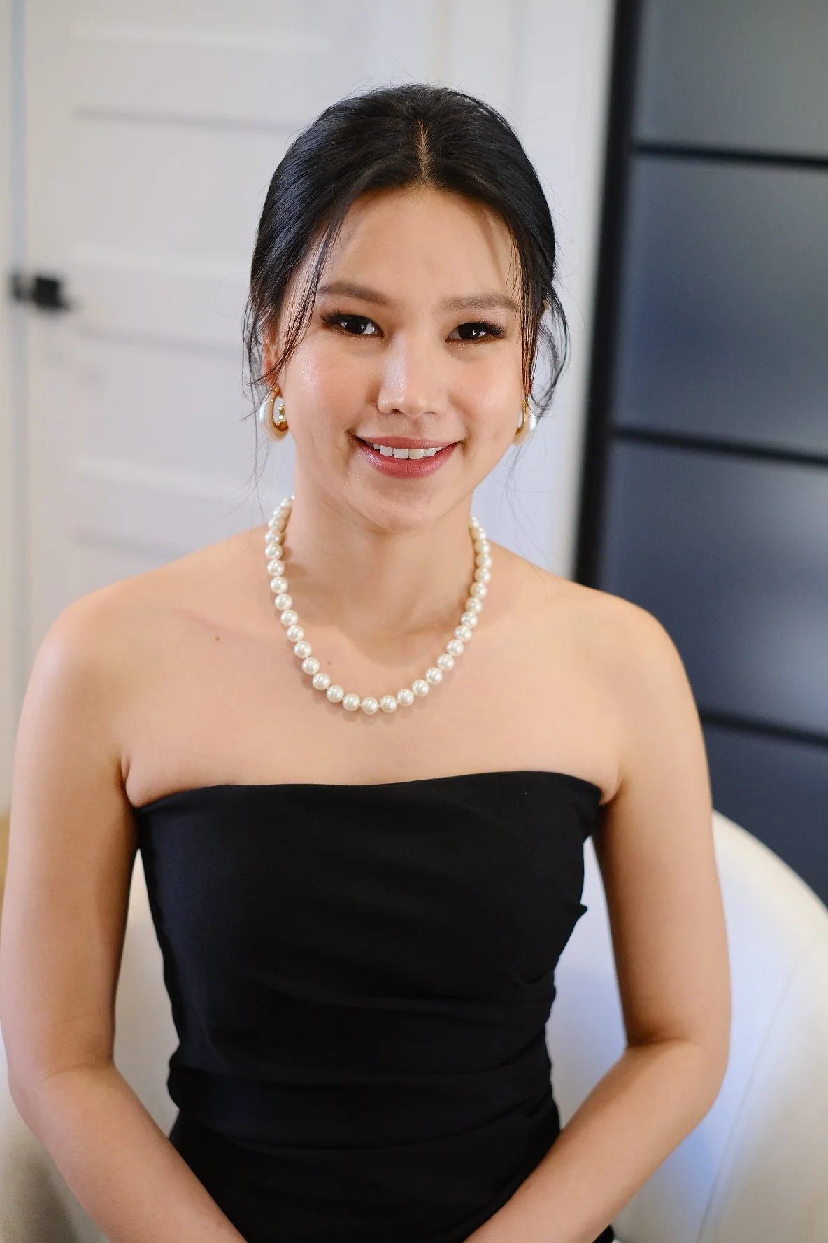 A woman with black hair styled in a bun, wearing a pearl necklace, pearl earrings, and a strapless black dress, smiling at the camera in an indoor setting.