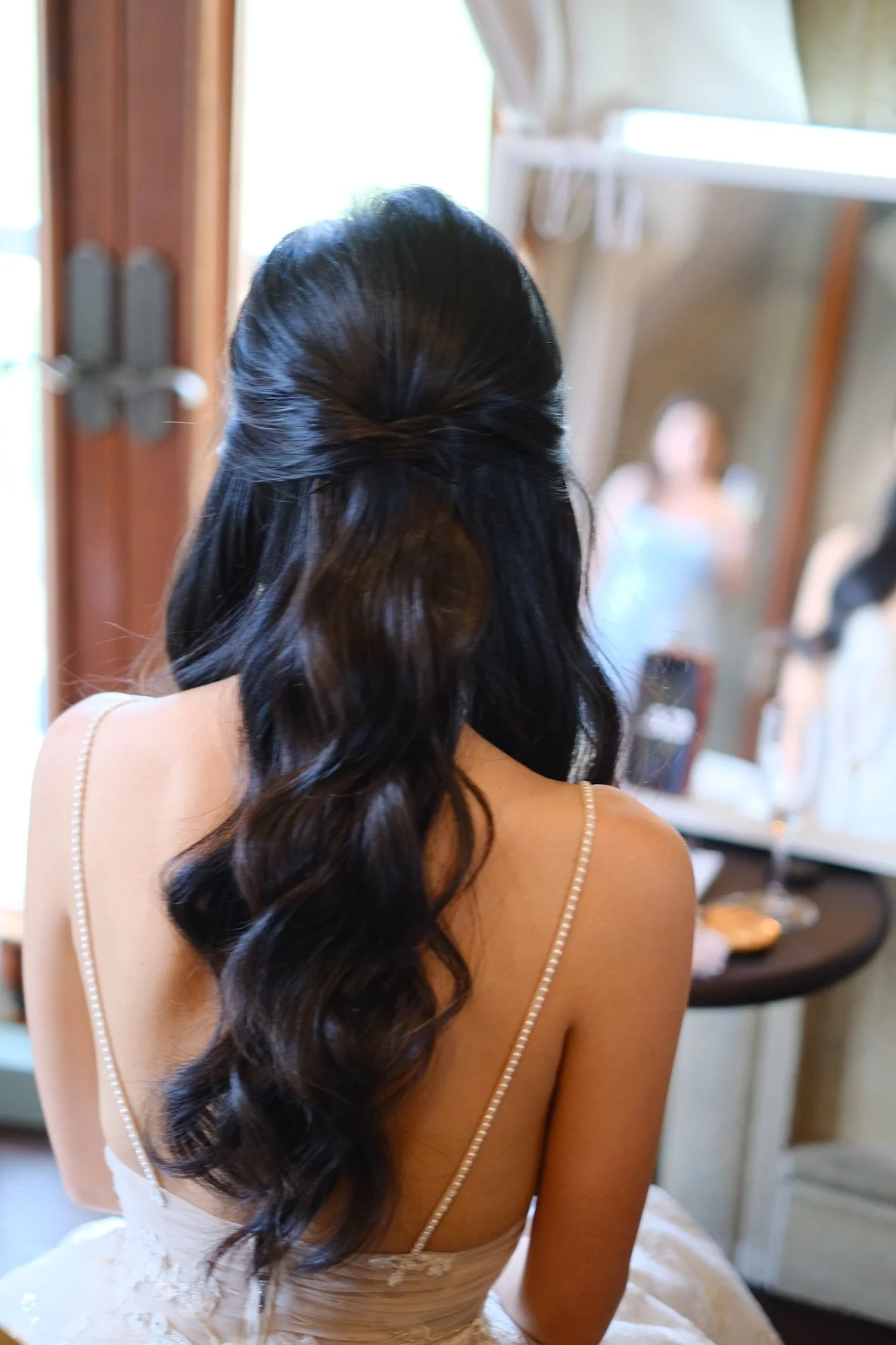 Back of a woman with long, wavy dark hair in a loose braid, wearing a beige dress with thin straps, possibly at a wedding or formal event, looking into a mirror with a woman in the background.
