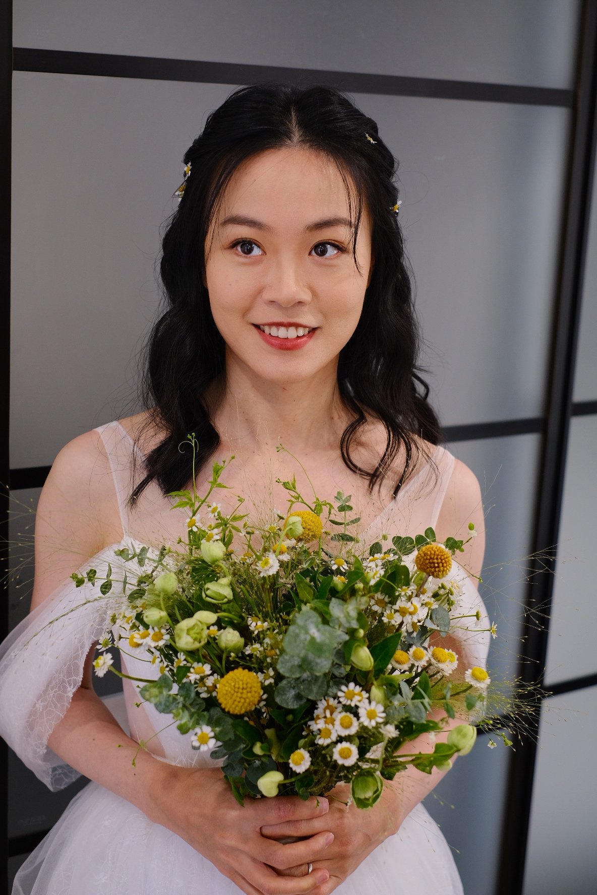 A woman with dark wavy hair and a white dress holding a bouquet of yellow and white flowers, smiling, with a gray background.
