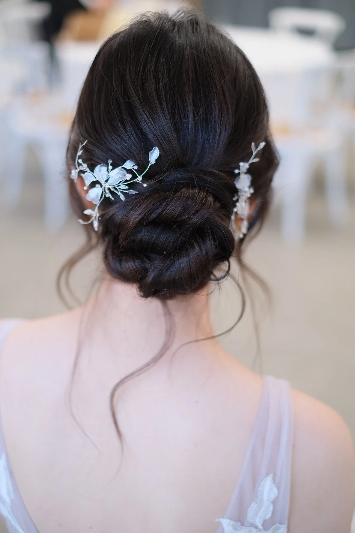 Back of a woman with dark hair styled in an elegant updo, decorated with white floral hairpieces, wearing a white dress with sheer fabric and floral appliqué details.