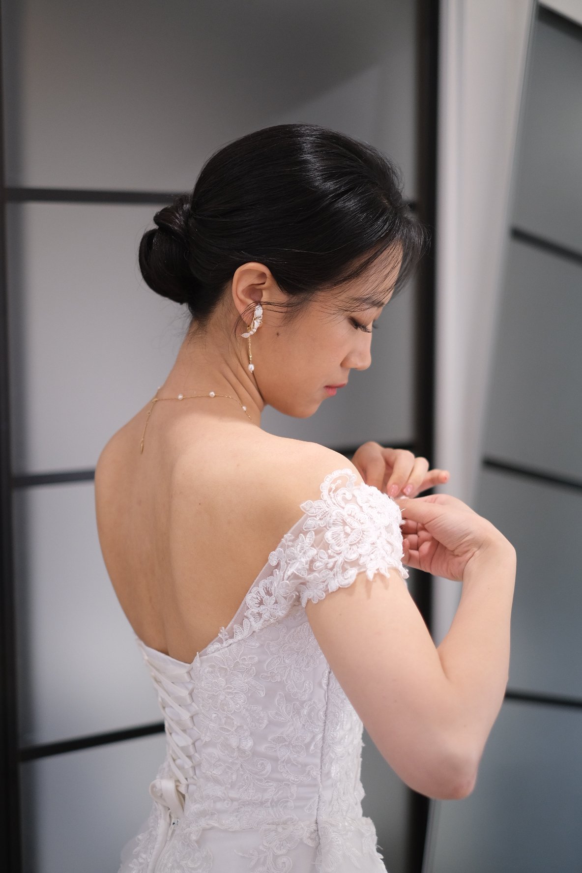 Bride with black hair styled in an elegant updo, wearing lace wedding dress with floral details, and jewelry, adjusting her dress or accessory.