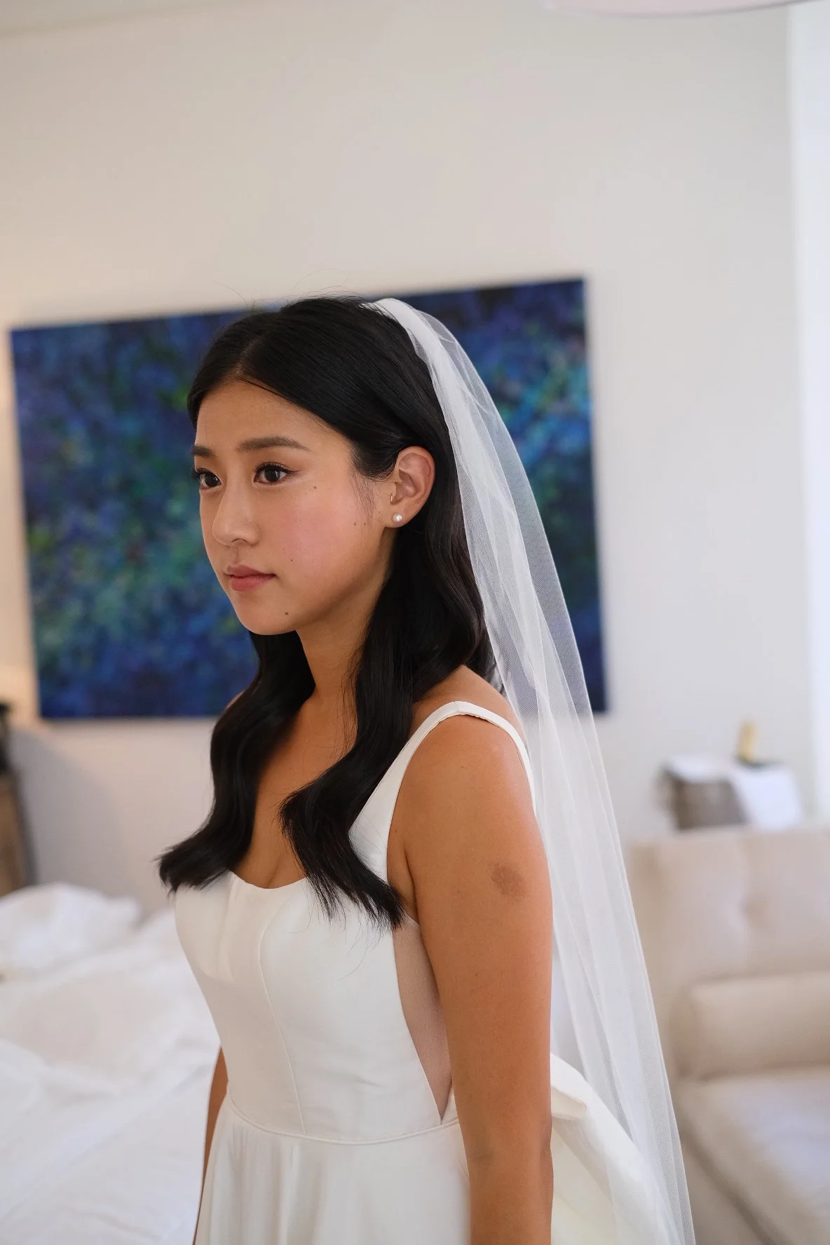 A young woman with dark hair and pearl earrings in a white wedding dress and veil standing indoors.