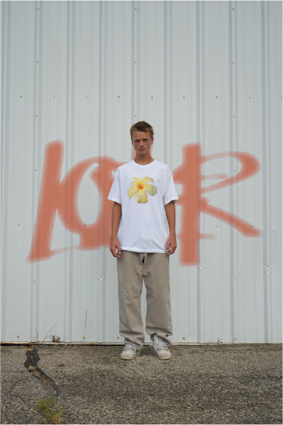 Young man standing in front of a white metal wall with red graffiti spelling 'LOVER' and a yellow flower graphic on his white t-shirt, wearing beige pants and sneakers.