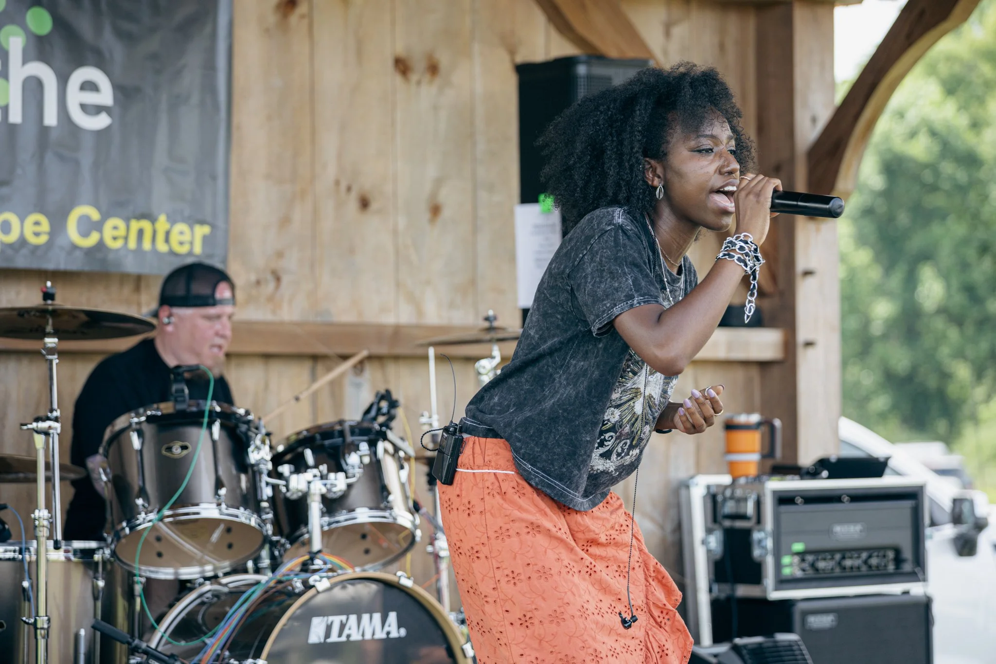 A woman with curly hair singing into a microphone during a live performance, with a drummer playing drums in the background inside a wooden pavilion with natural light.