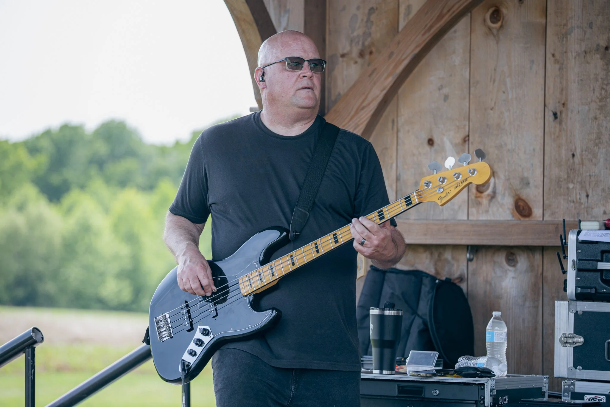 A man wearing sunglasses and a black t-shirt playing an electric bass guitar outdoors.