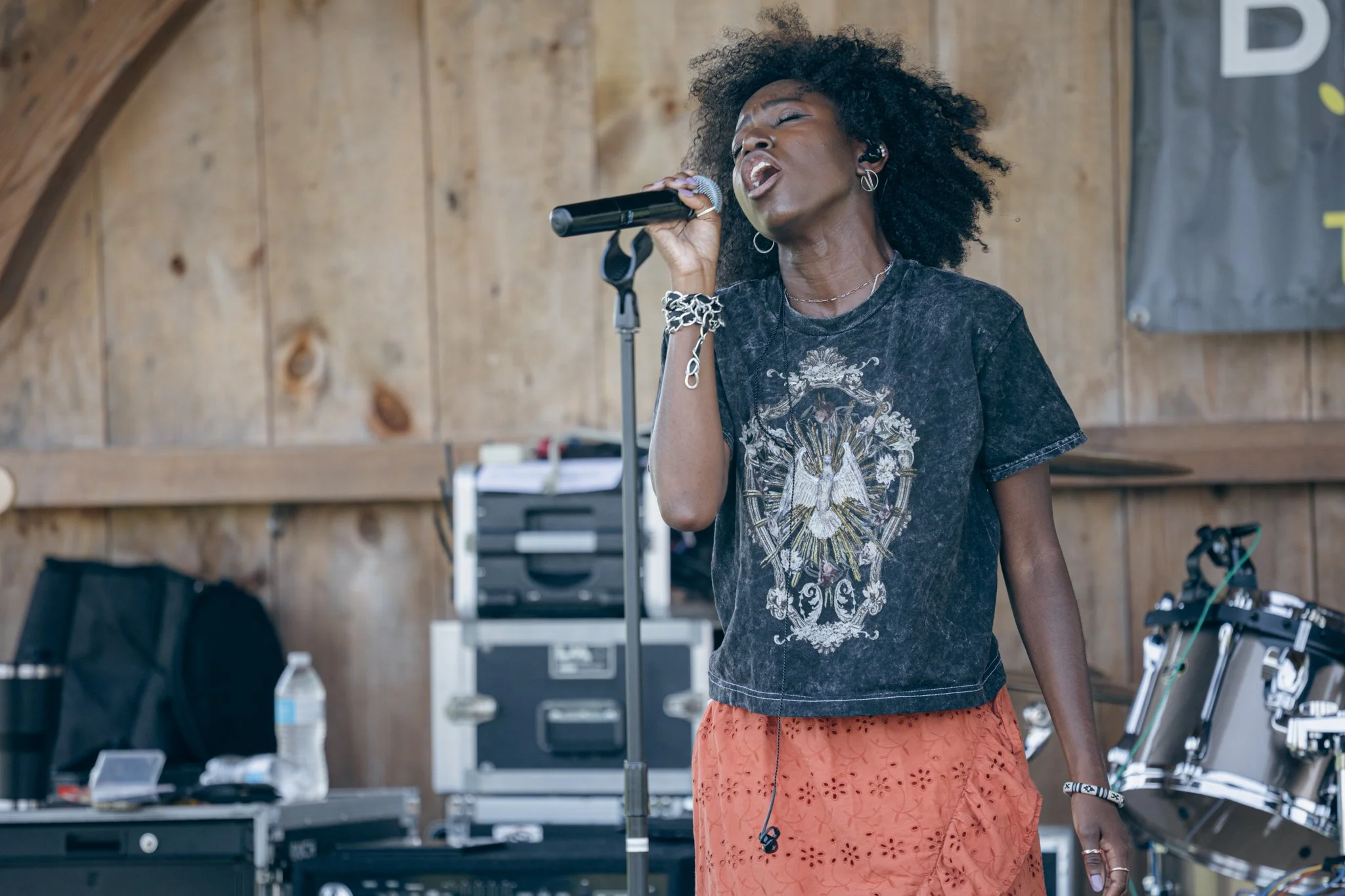 A woman singing passionately into a microphone with her eyes closed, wearing a dark t-shirt with a colorful graphic, jewelry including earrings and bracelets, and an orange skirt, in a rustic setting with wood paneling and musical equipment visible i