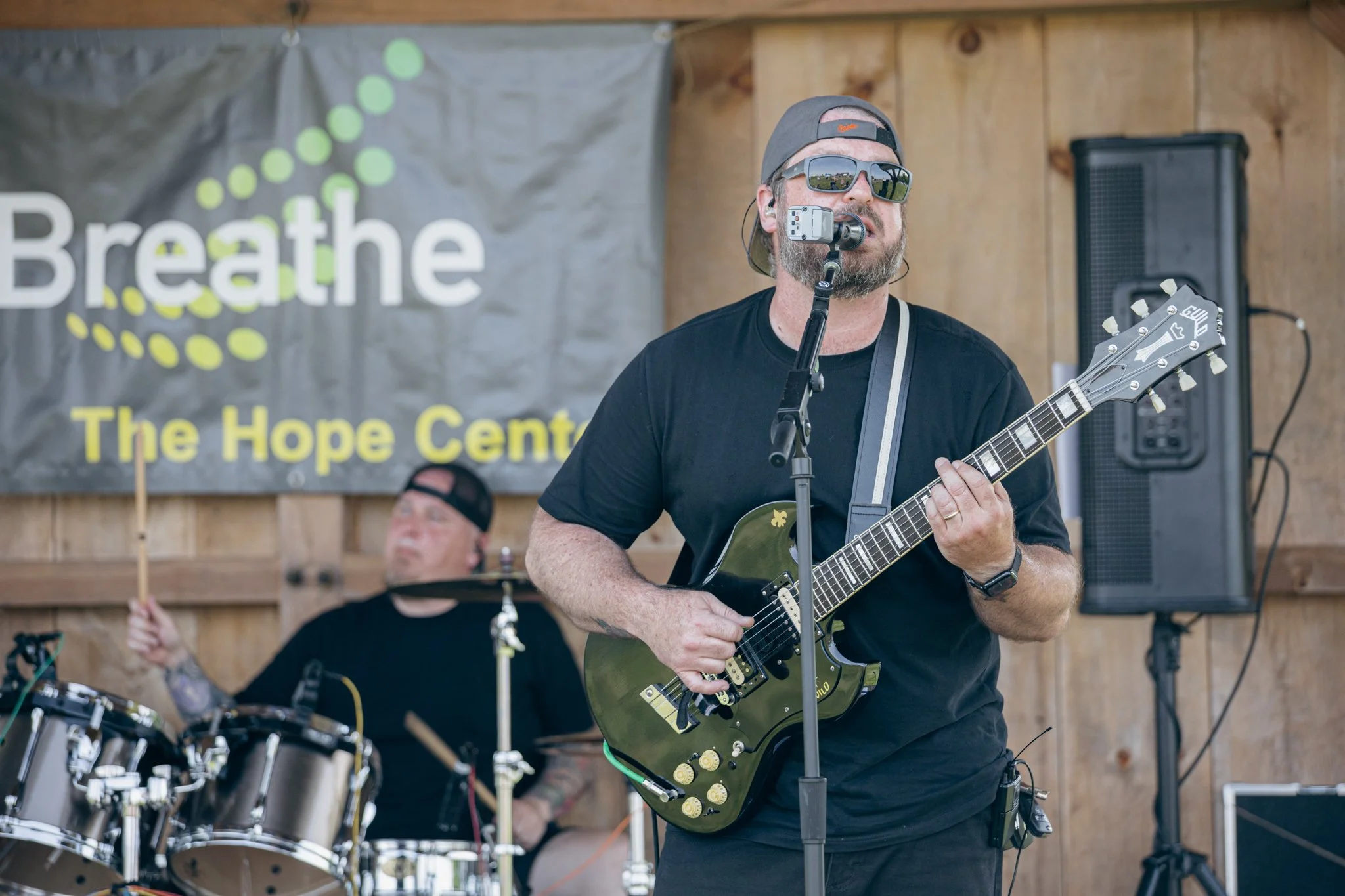 A man with a beard wearing sunglasses, a backwards cap, and a black shirt playing an electric guitar during a live performance. In the background, a drummer with tattoos and a black cap plays drums. There is a banner that reads 'Breathe The Hope Cent