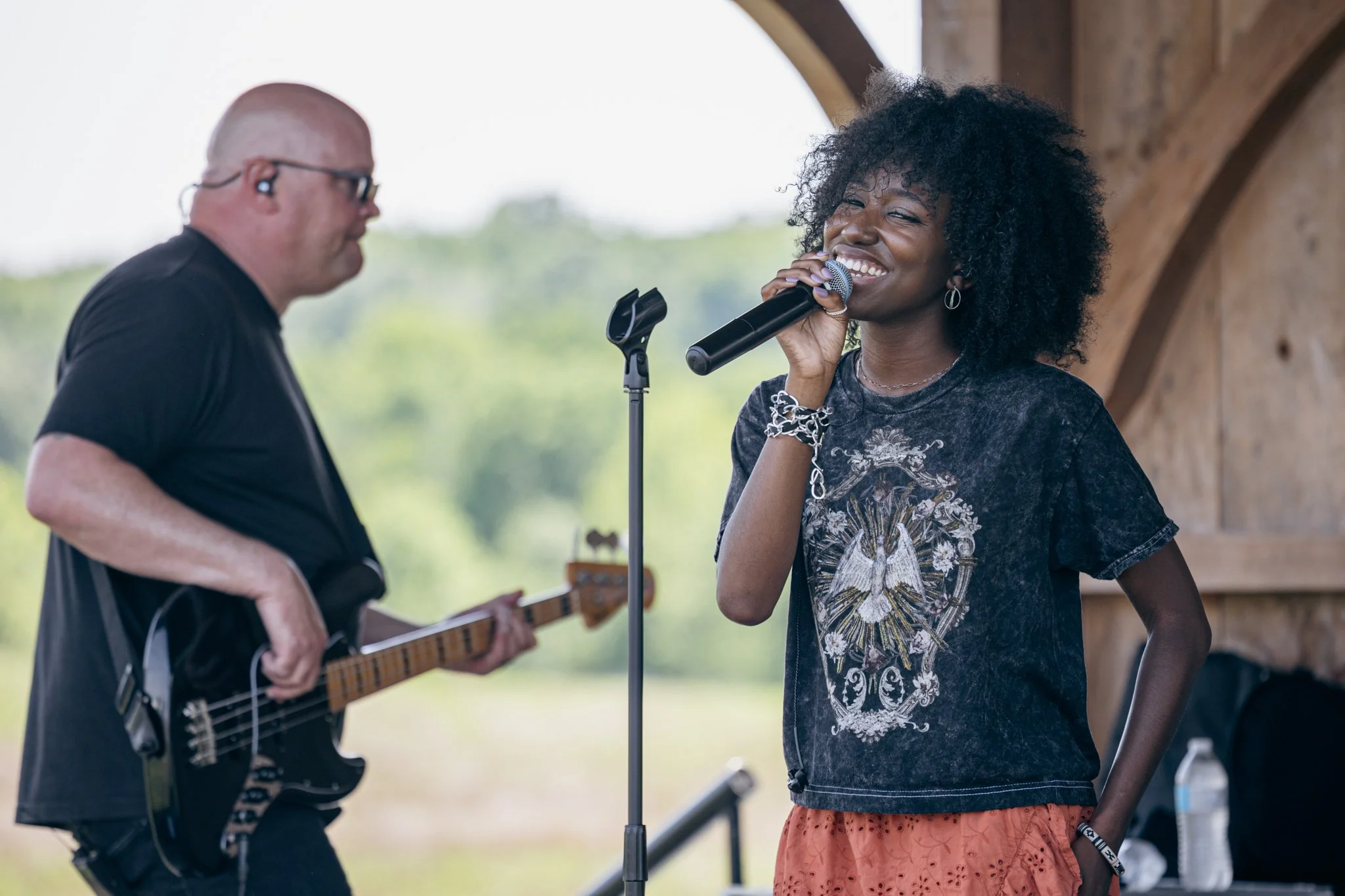 A woman with curly hair singing into a microphone with a man in sunglasses playing guitar beside her at an outdoor event.