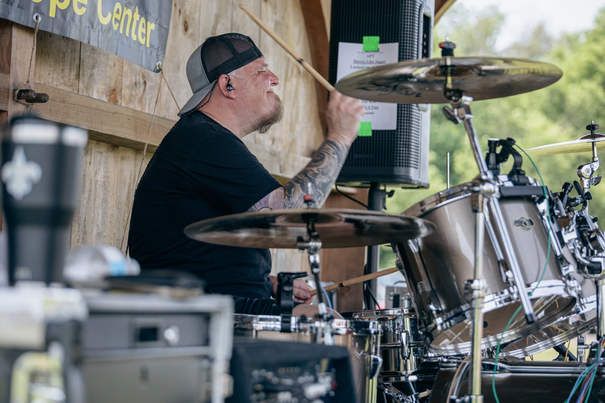 A man playing the drums at an outdoor event, wearing a black baseball cap backwards and ear monitors, with trees in the background and a wooden wall behind him.