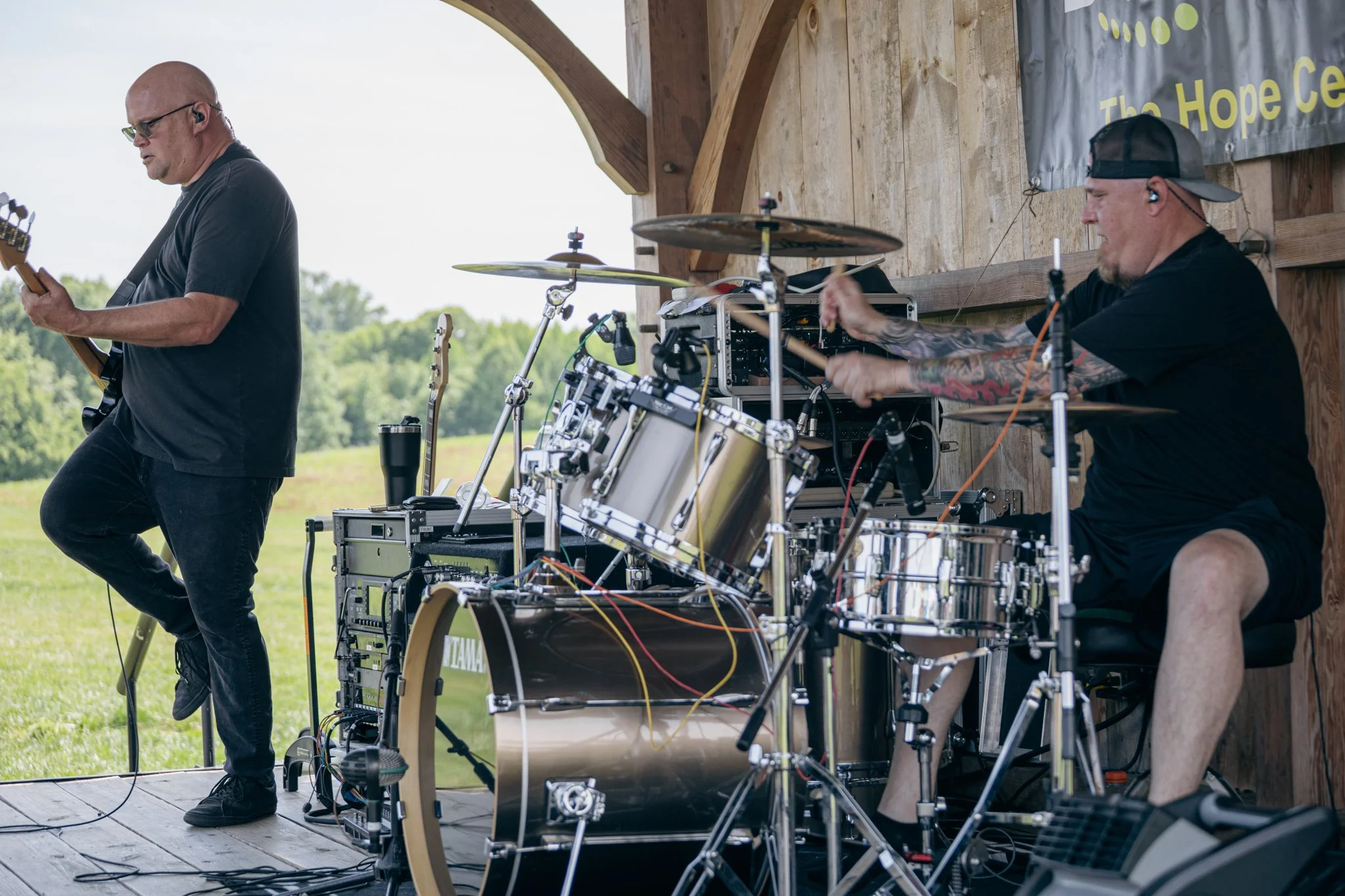 Two musicians performing outdoors on a wooden stage, one playing guitar and the other on electronic drum set, with a banner behind them reading 'The Hope Ce'.