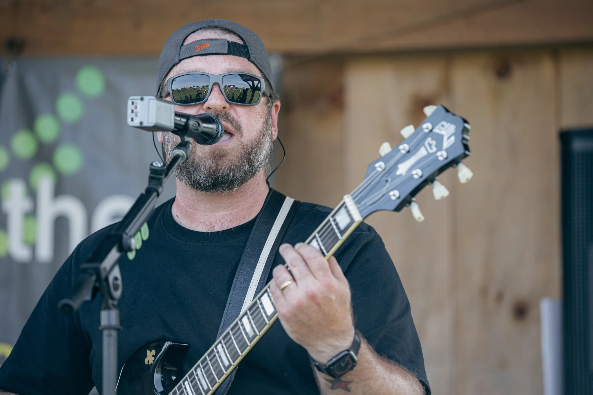 A man wearing sunglasses and a baseball cap, singing into a microphone while playing an electric guitar, at an outdoor event.