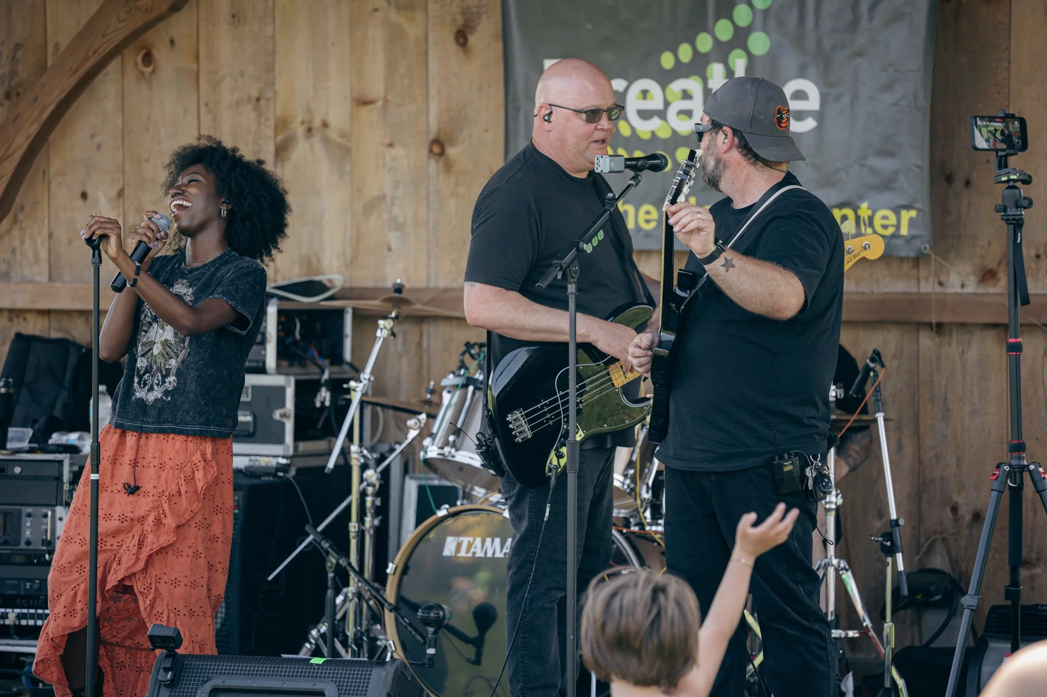 A woman with curly black hair singing into a microphone, a man in sunglasses and a black t-shirt playing a guitar, and another man in a cap holding a guitar, performing on a wooden stage with musical equipment.