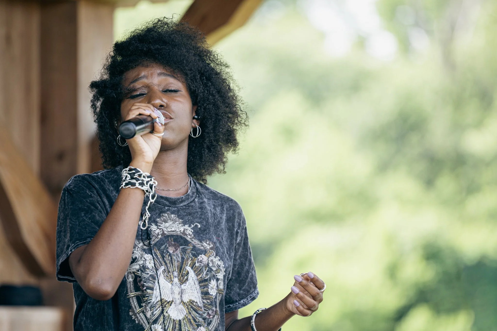 A woman with curly black hair and earrings singing into a microphone outdoors.
