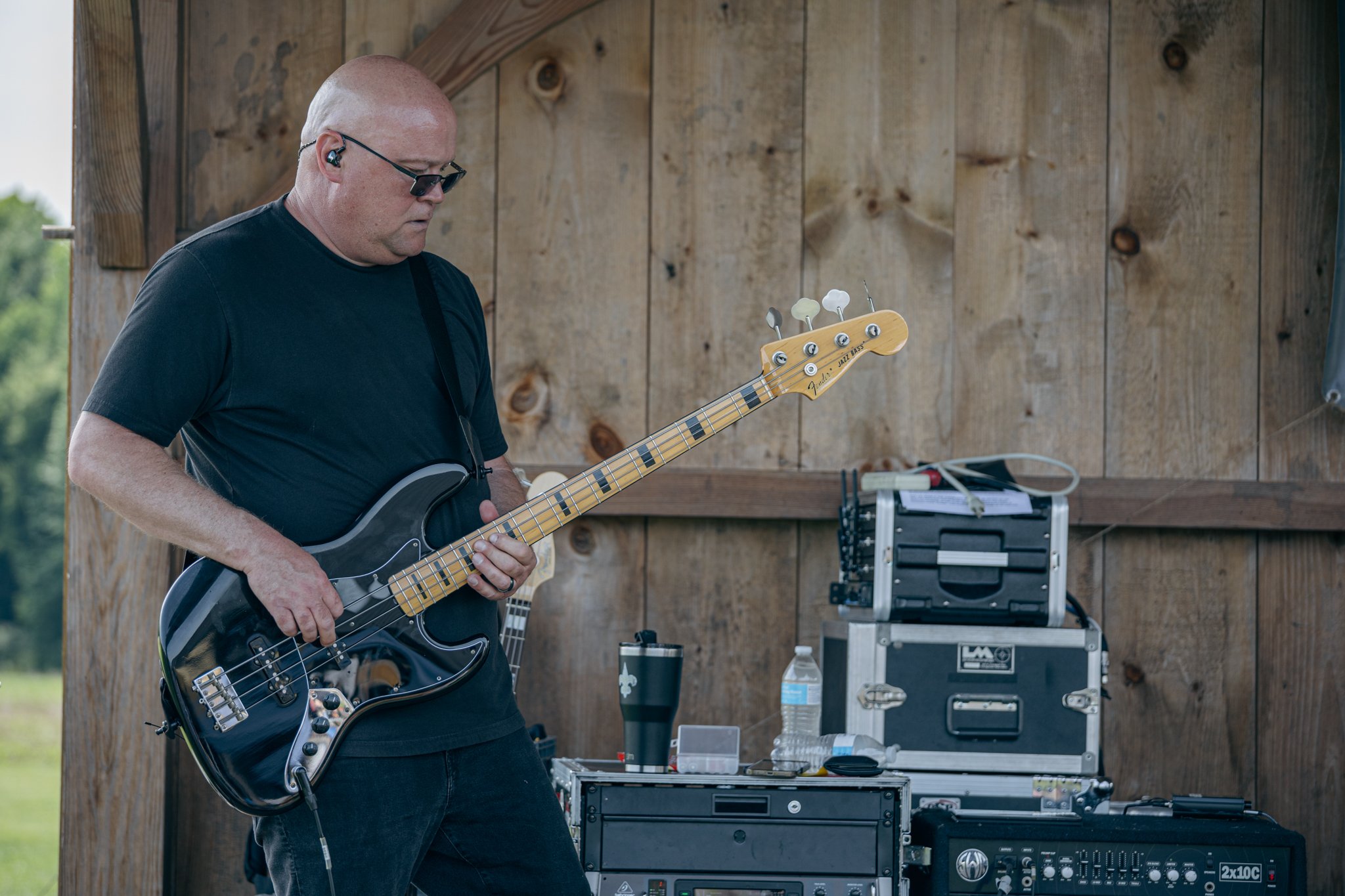 A bald man wearing glasses and earbuds playing a black electric bass guitar outdoors in front of a wooden wall, with music equipment and a water bottle nearby.
