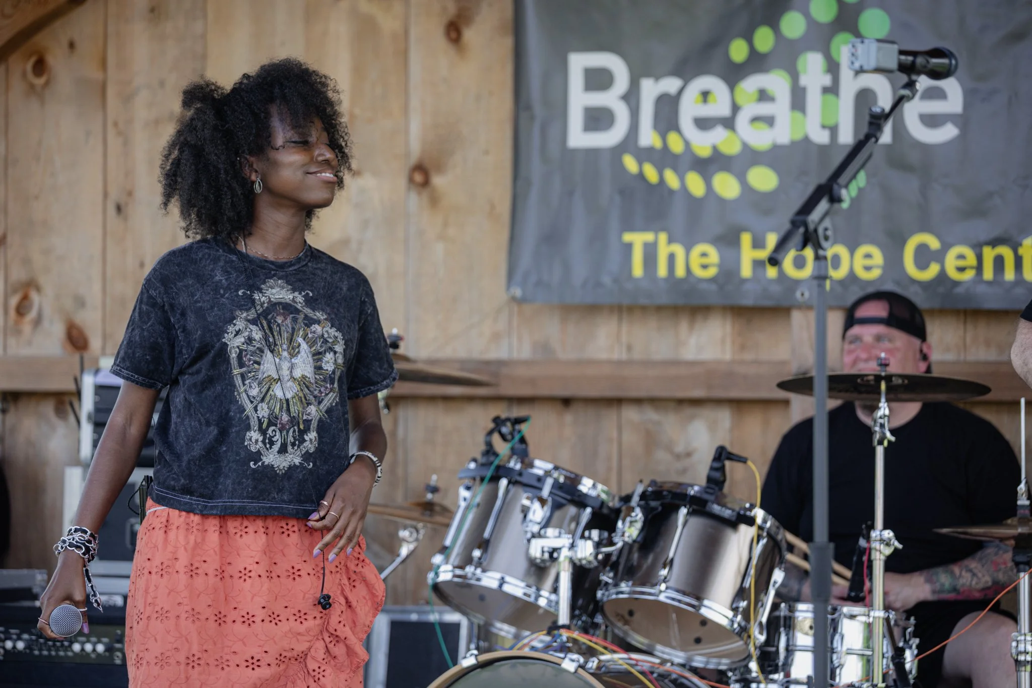 A woman with curly black hair smiling, holding a microphone, standing in front of a drummer and musical equipment at an indoor event with a wooden wall and a banner that reads 'Breathé The Hope Cent'.
