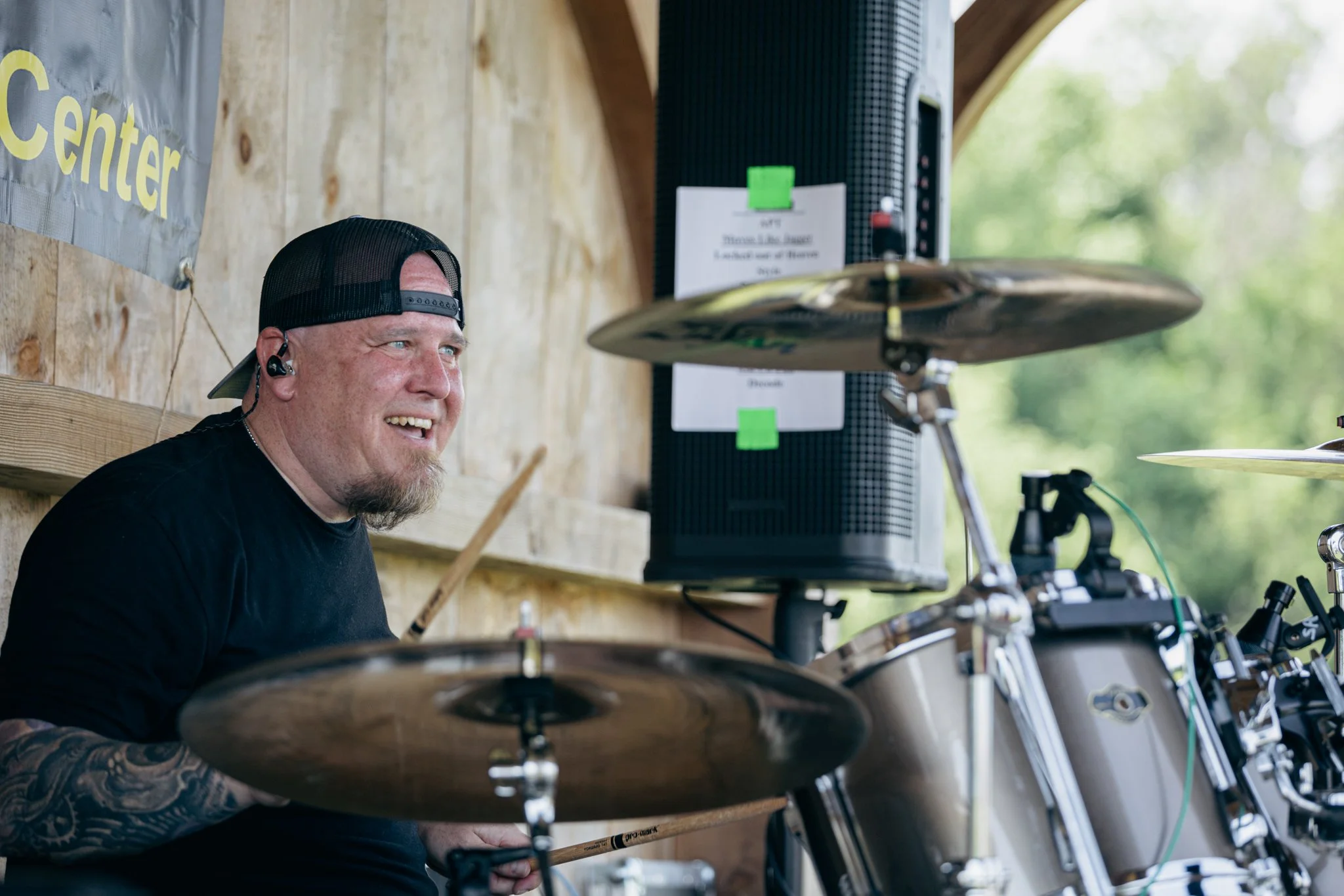 A smiling man with a tattooed arm, wearing a backward black cap and in-ear monitor, playing drums outdoors during daytime.
