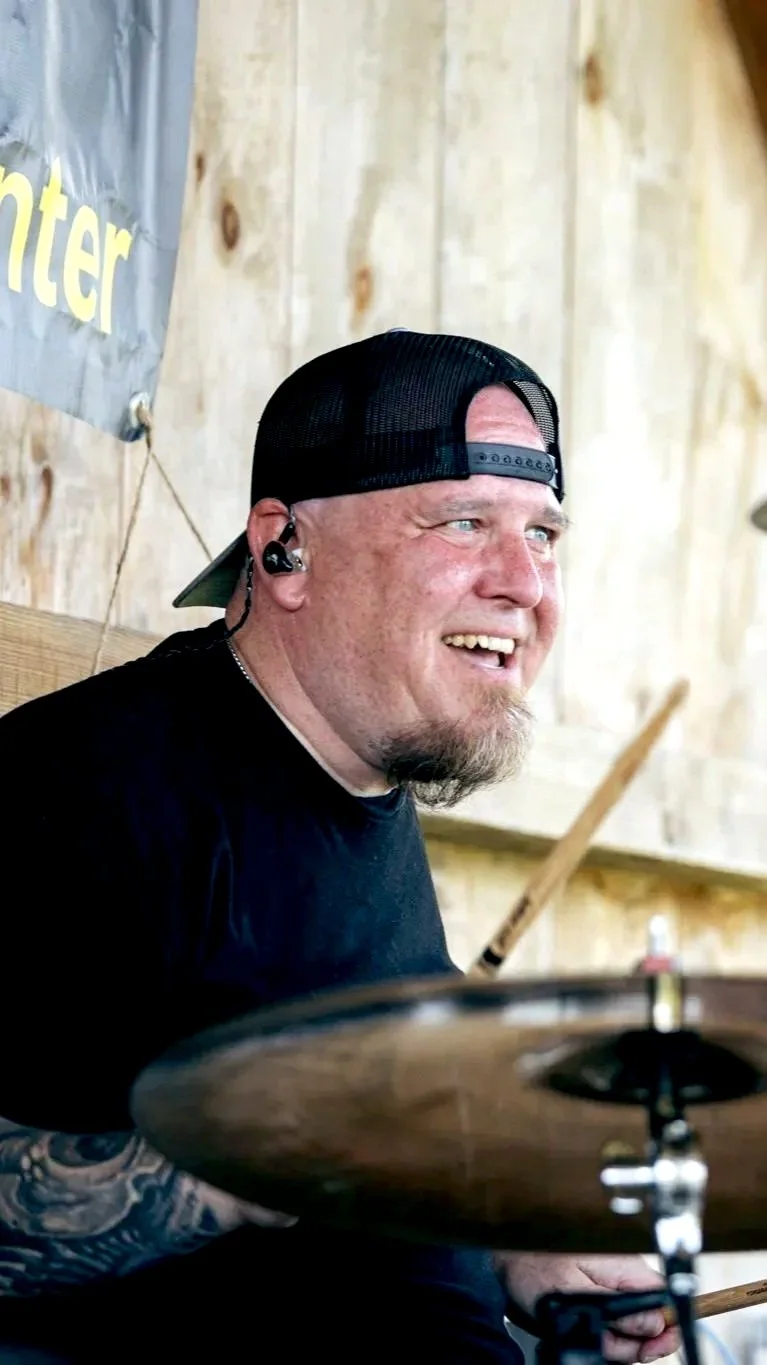 A smiling man wearing a black baseball cap backwards, black T-shirt, and listening to in-ear monitors, sitting in front of a drum set, with a wooden background and part of a gray sign visible.