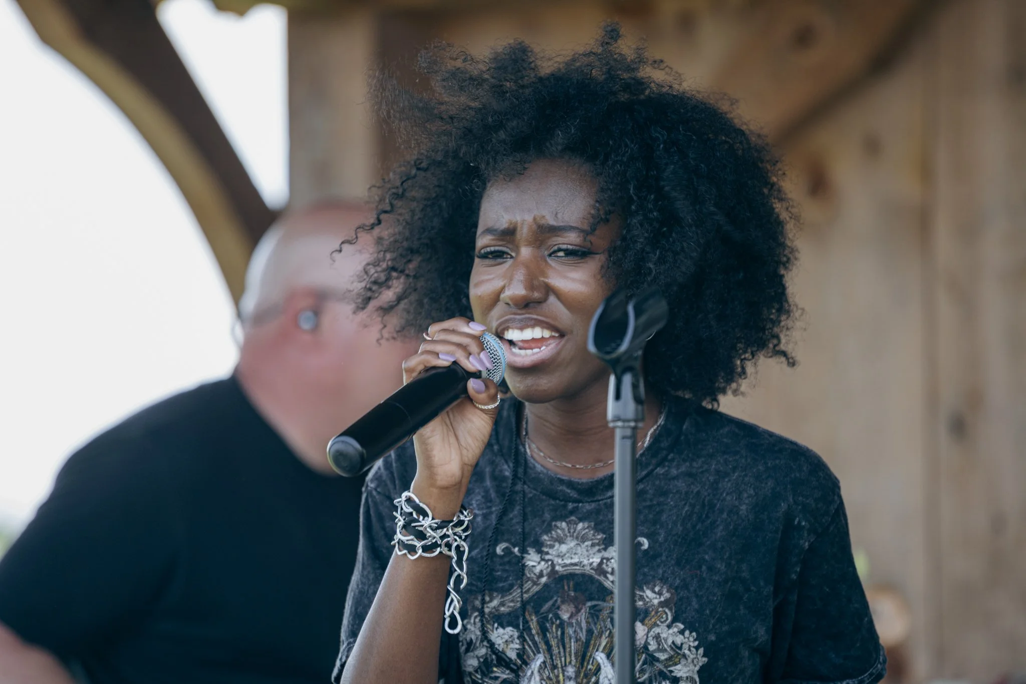 A woman with curly black hair singing into a microphone held by a person in front of her, outdoors, with a wooden structure in the background.