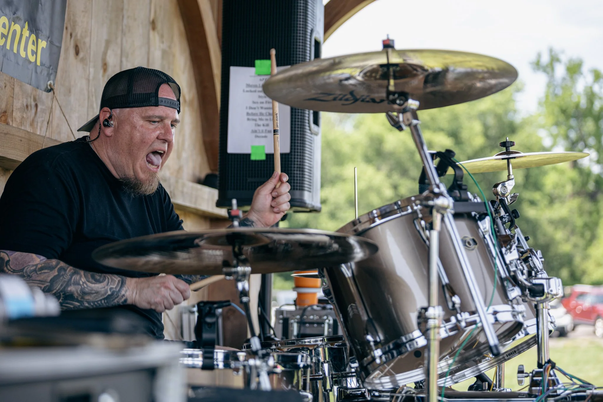 A man wearing a black cap backwards, black T-shirt, and earbud, intensely playing drums at an outdoor setting with a wooden backdrop and greenery in the background.