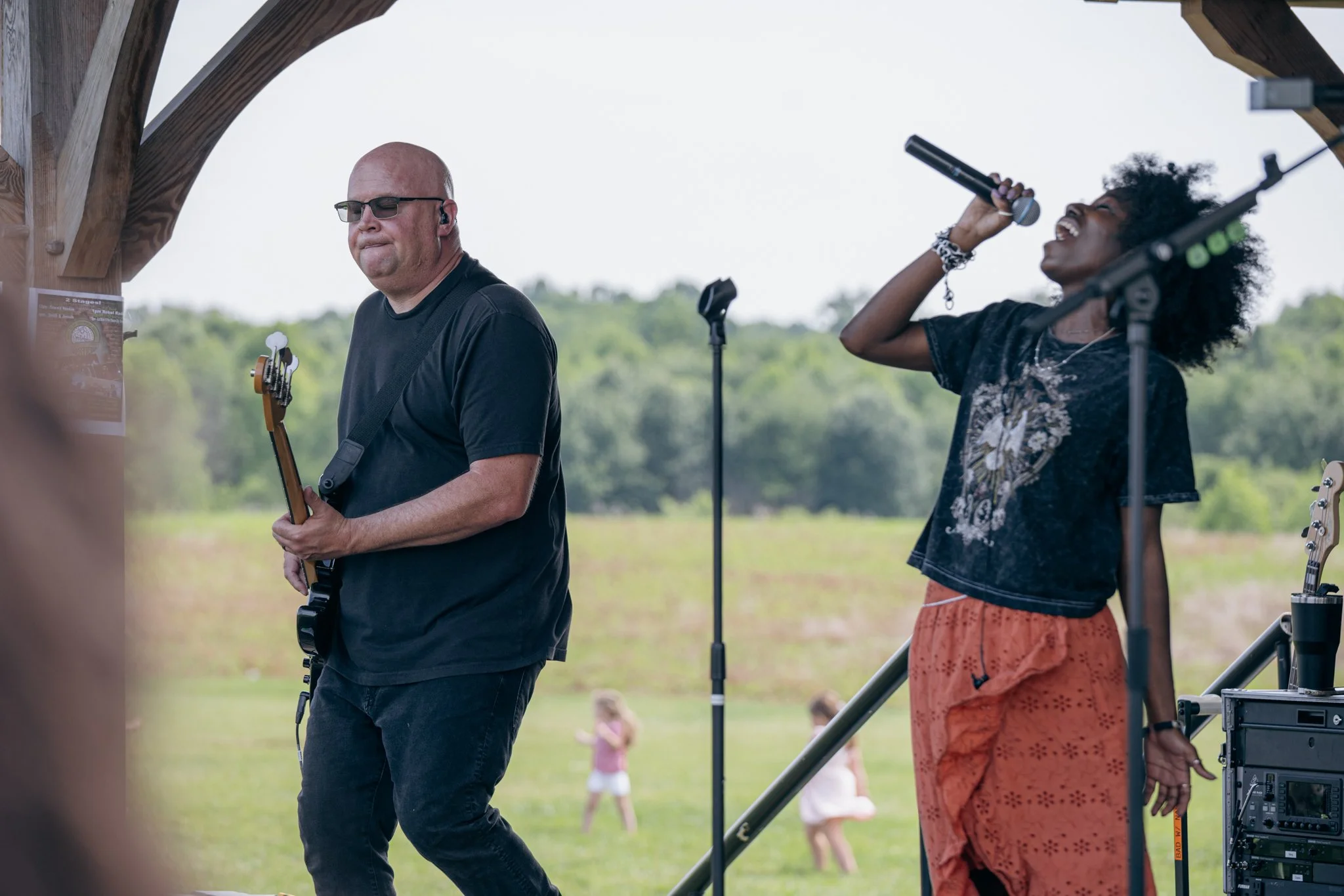 Two musicians performing outdoors under a wooden pavilion. The male musician on the left is playing an electric guitar, wearing glasses and a black shirt. The female musician on the right is singing into a microphone, with curly hair, wearing a patte