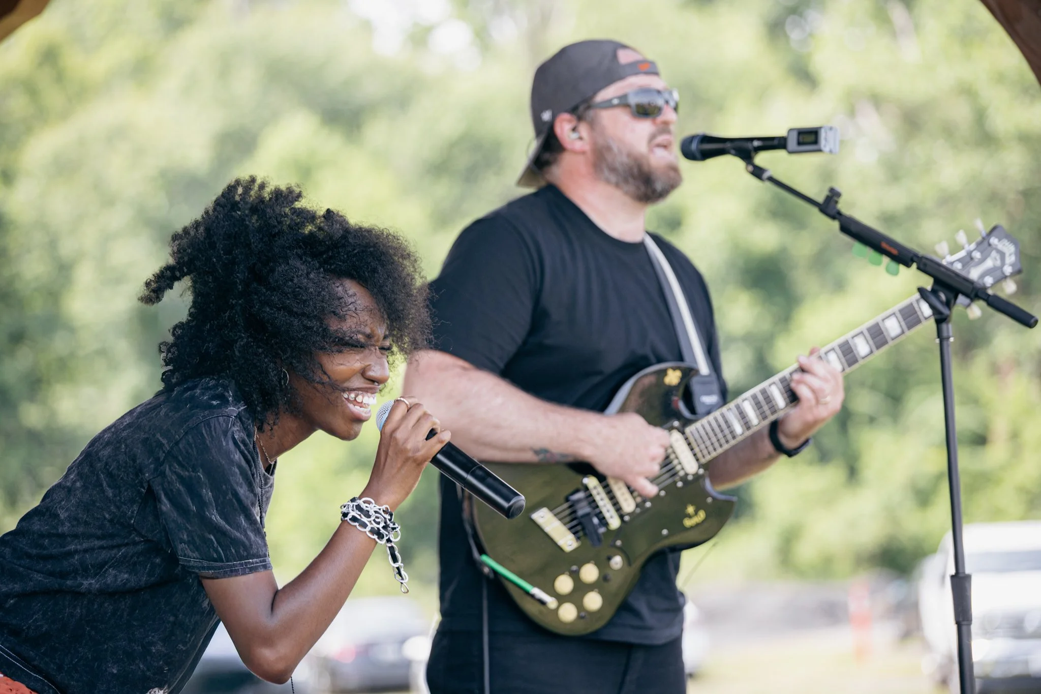 A woman singing into a microphone and a man playing an electric guitar outdoors at a park, with trees and bright natural light in the background.