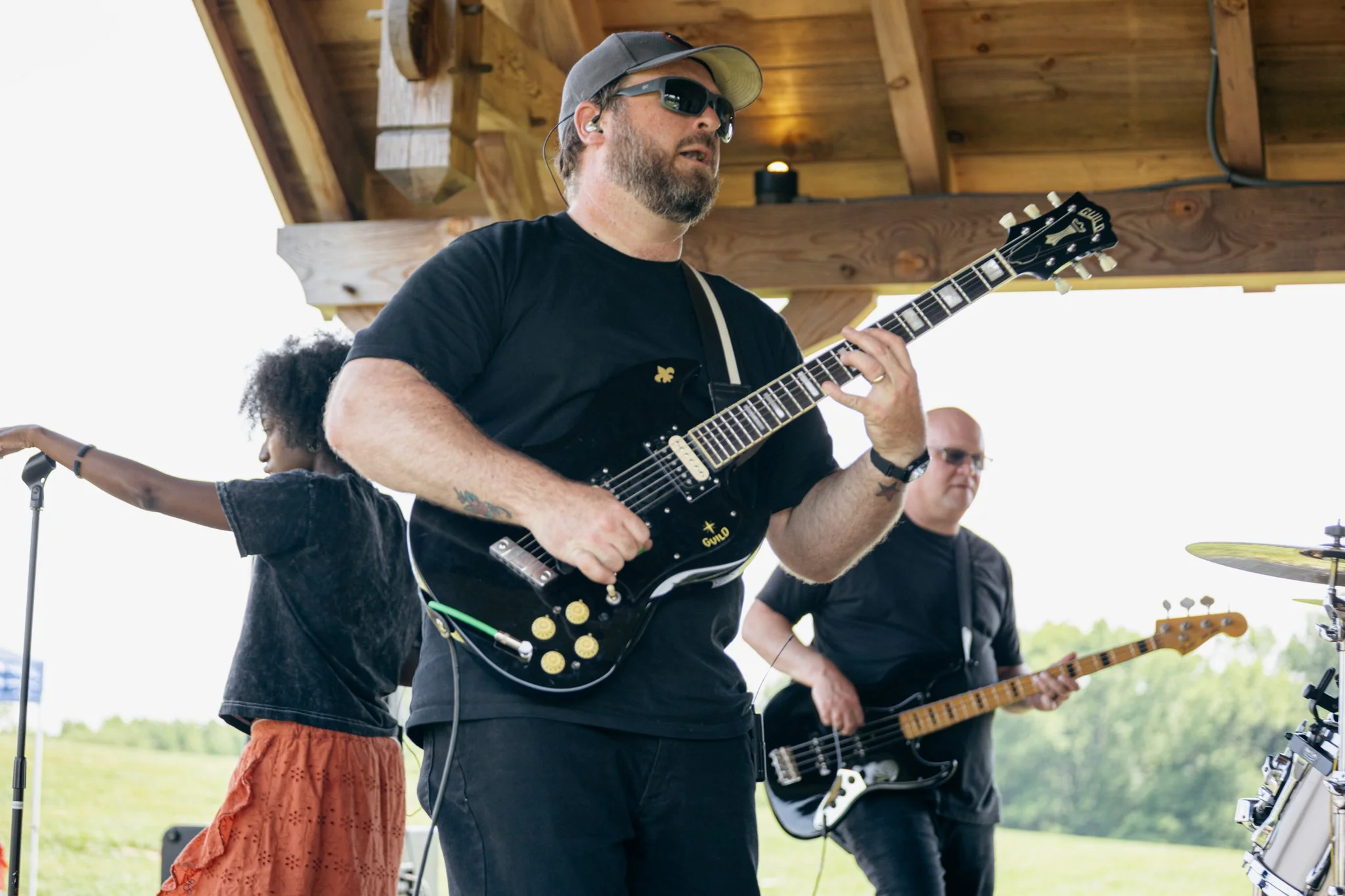 Musician playing guitar at an outdoor event, with a man holding bass guitar, a woman with curly hair, and a drum set partially visible.