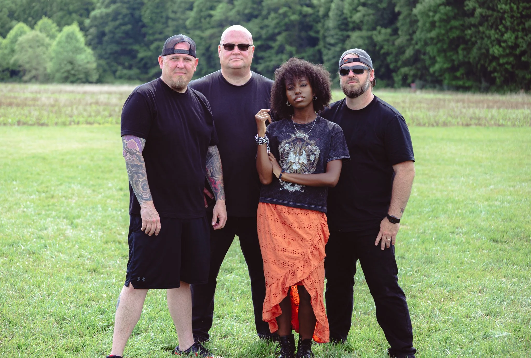 A group of four diverse people standing outdoors on a grassy field, framed by green trees in the background.