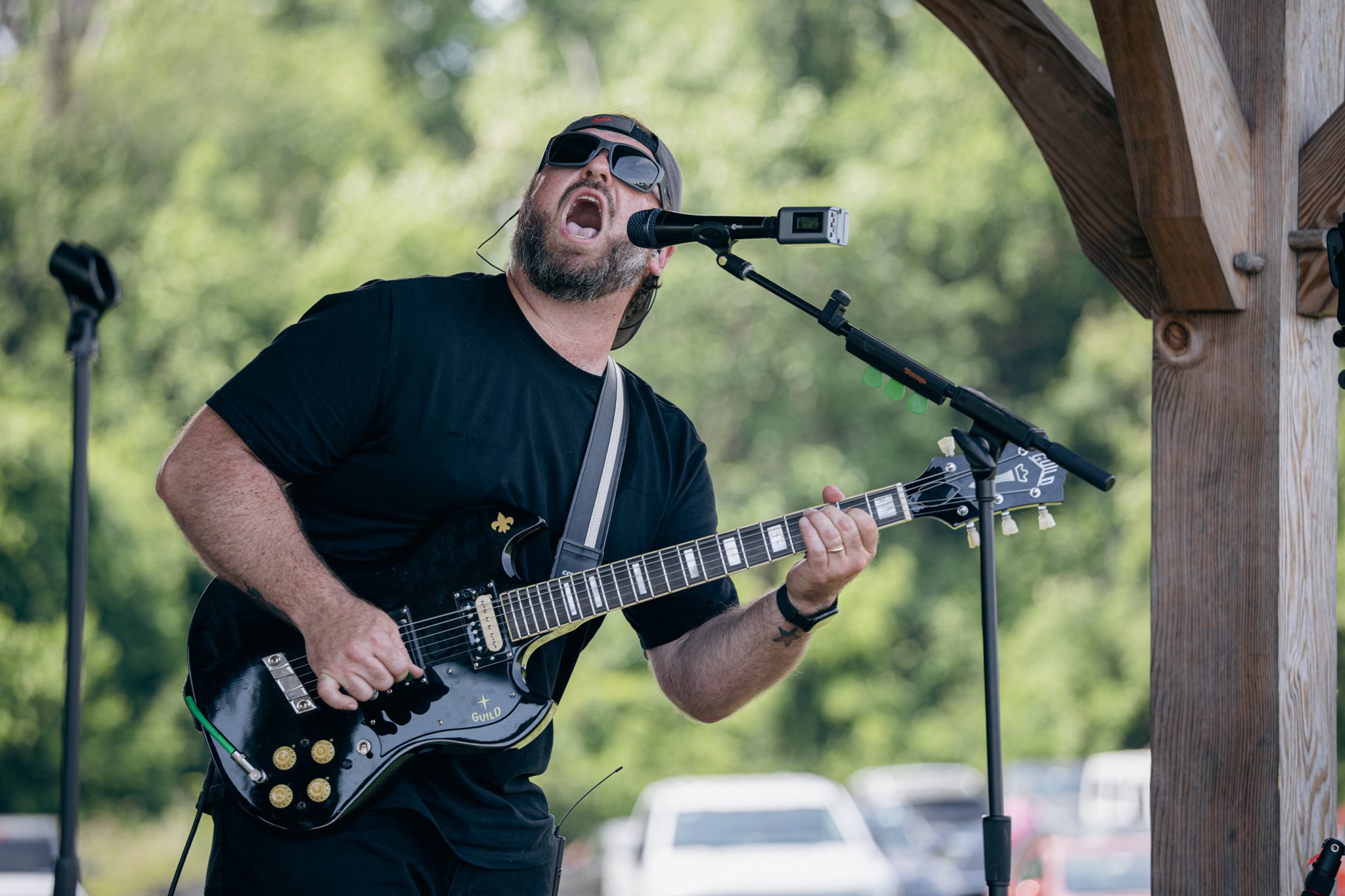 A man with a beard and sunglasses singing into a microphone while playing an electric guitar outdoors under a wooden structure, with green trees in the background.