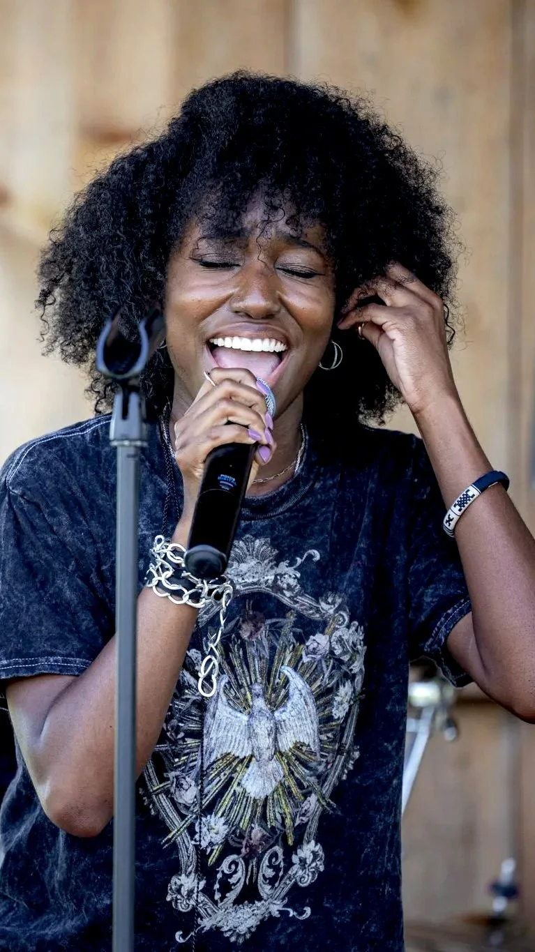A woman with curly hair singing into a microphone, wearing a black T-shirt with a detailed graphic design, and smiling with her eyes closed.