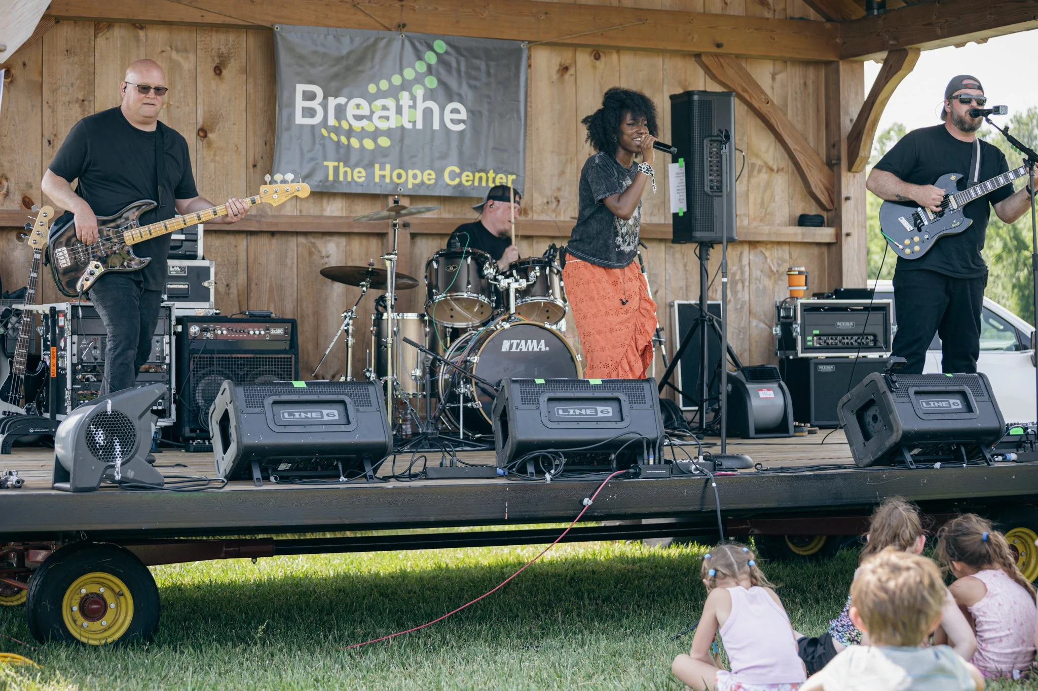 A band performing on an outdoor stage with children sitting on the grass in front. The stage has a wooden backdrop with a banner reading 'Breathe The Hope Center.' The band includes a guitarist, a drummer, and a female vocalist singing into a microph