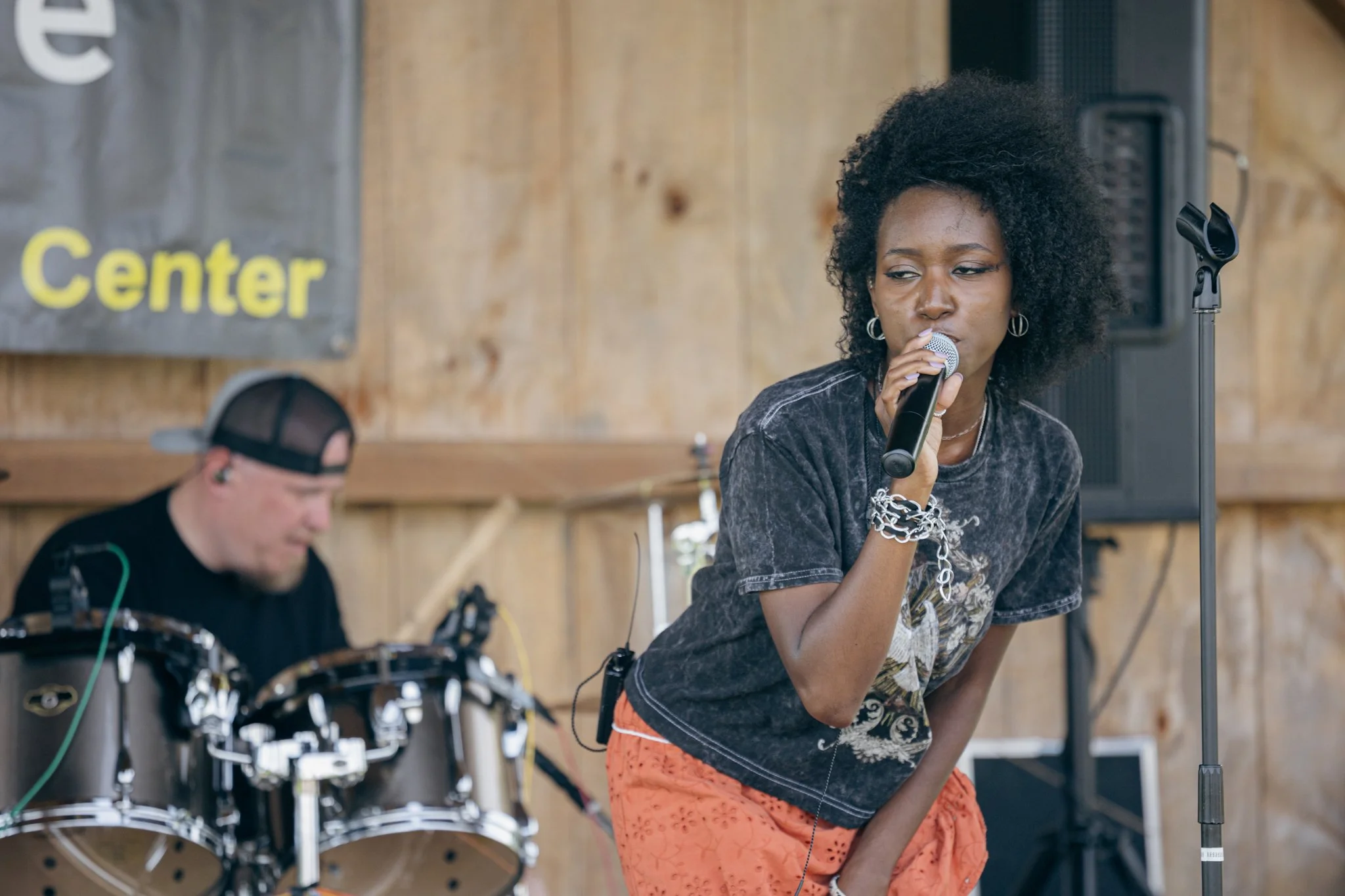A woman singing into a microphone during a live performance, with a drummer in the background in a wooden-finished room or venue.