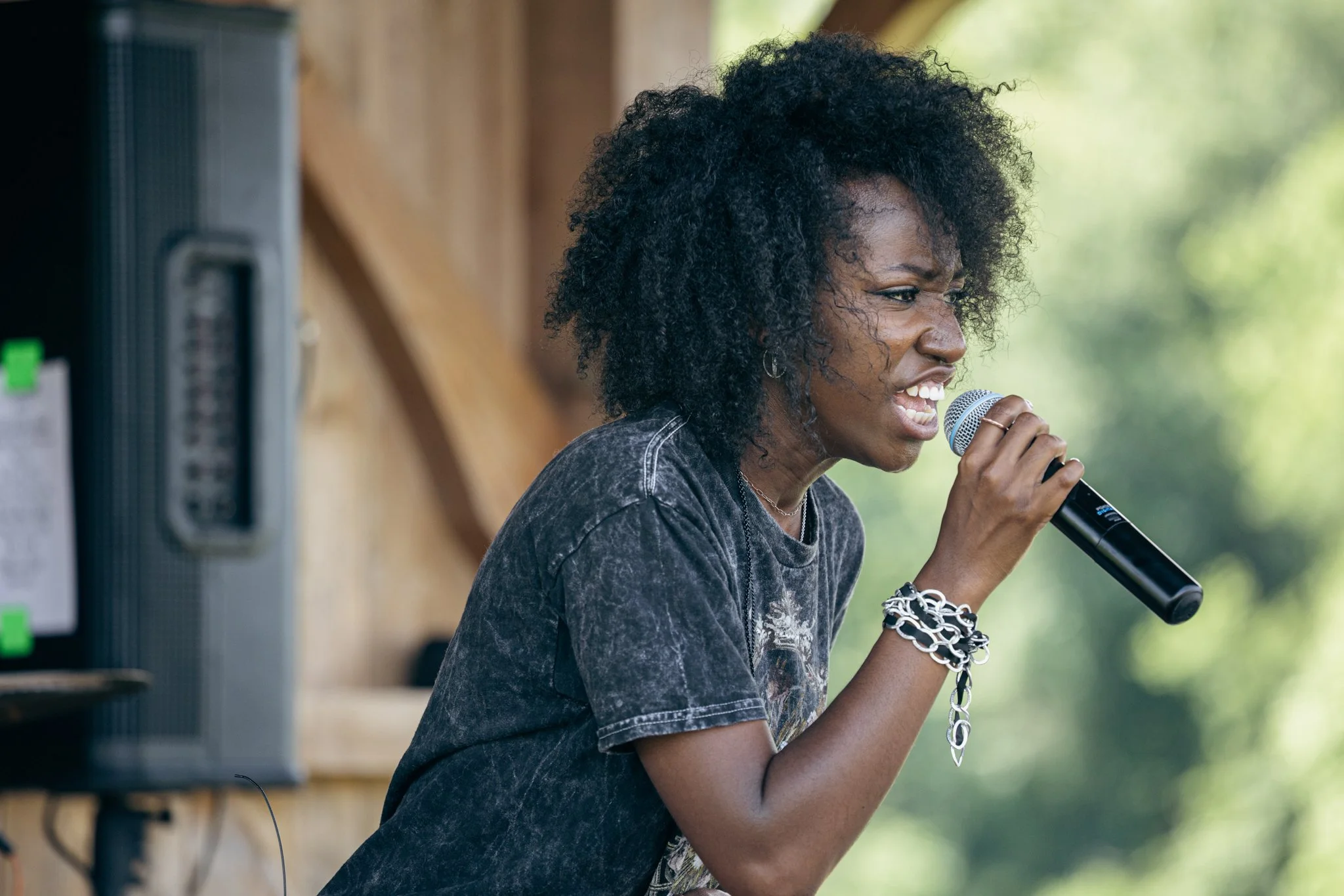 A woman with curly black hair singing into a microphone outdoors, wearing a black t-shirt and silver jewelry.