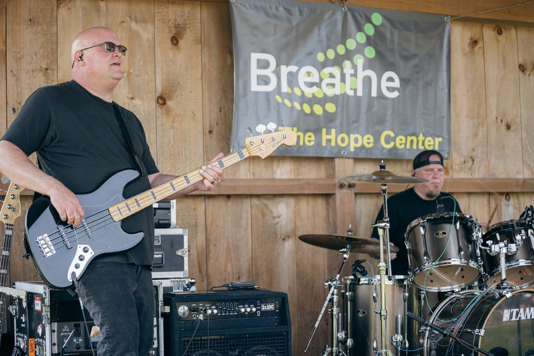 A man playing an electric bass guitar, wearing sunglasses and earbuds, stands in front of a wooden wall with a banner that reads 'Breathe The Hope Center'. Another man is sitting behind a drum set.