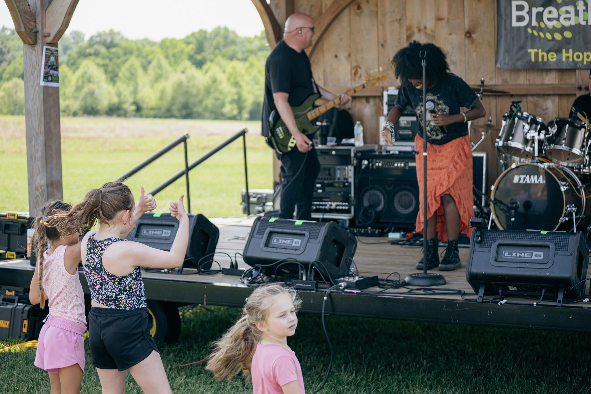 Children in front of a stage watching a band perform outdoors, with two girls giving thumbs up and one girl looking on, while musicians play instruments under a wooden canopy.