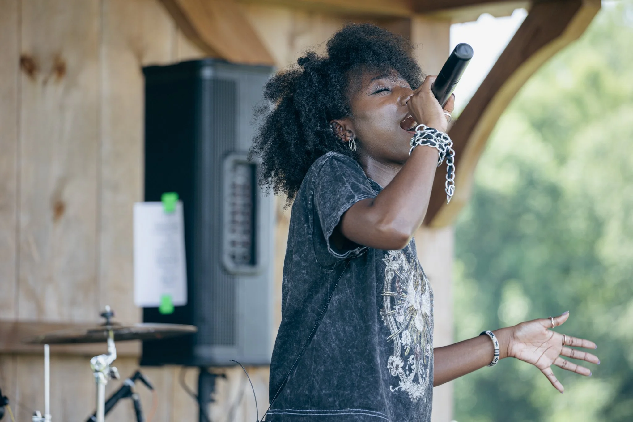 A woman with curly hair singing into a microphone indoors with a wooden background and large window showing greenery outside.