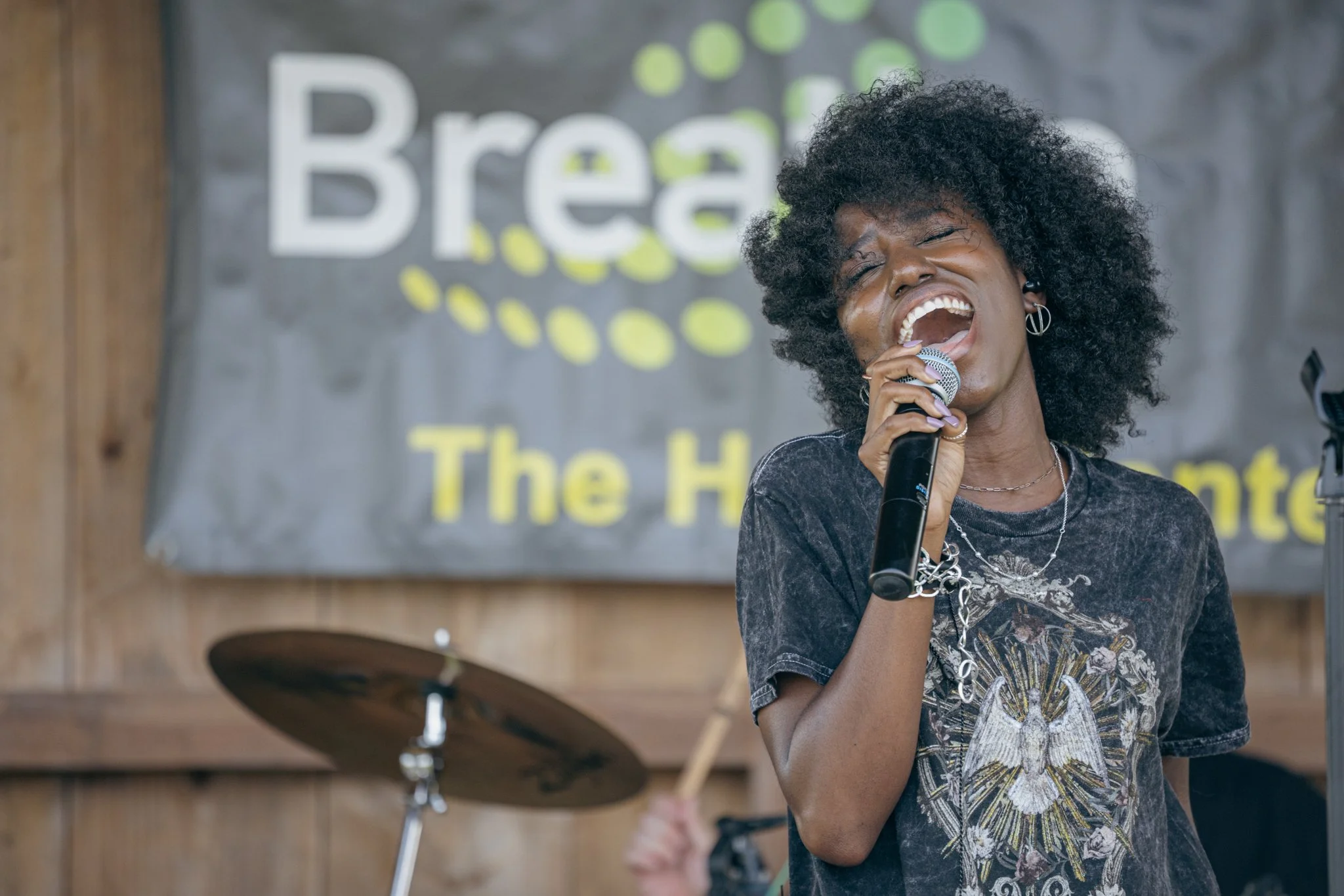 A woman singing into a microphone at an event with a sign in the background that reads 'The H'.