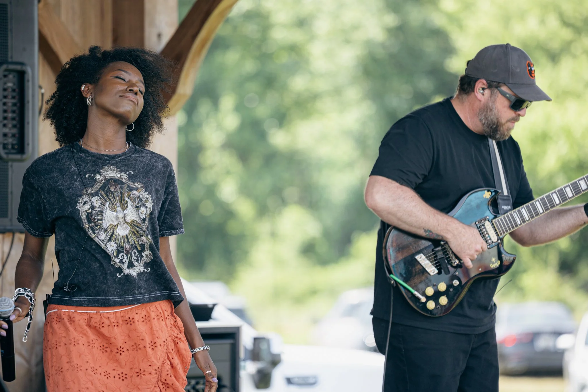 A woman with curly hair wearing a dark graphic t-shirt and orange skirt appears to be enjoying music with her eyes closed, standing next to a man playing electric guitar outdoors.