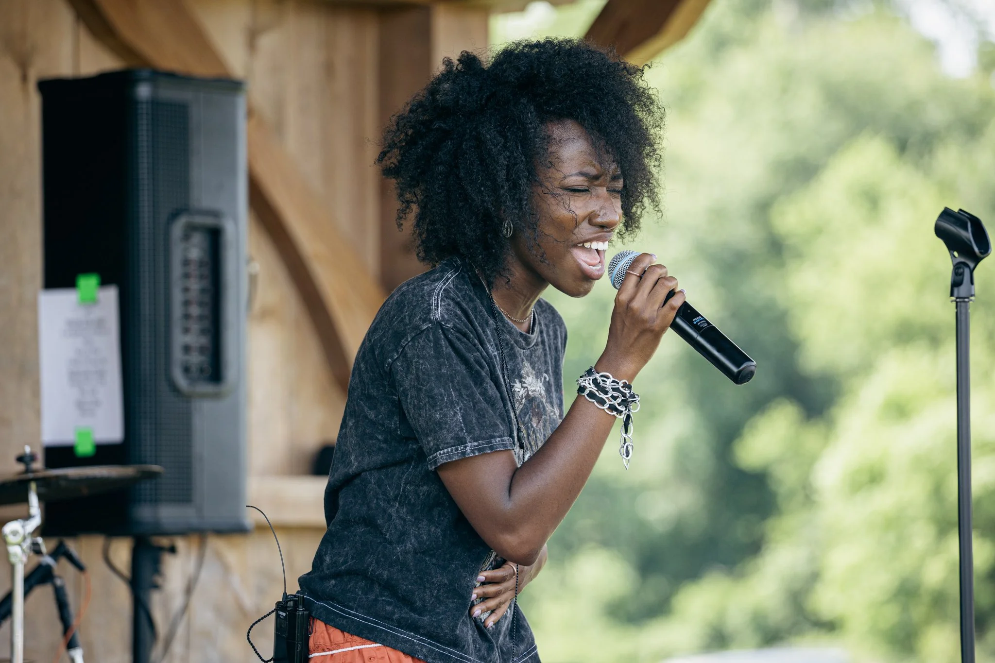 A woman with curly black hair singing into a microphone outdoors, wearing a dark t-shirt and jewelry, standing near sound equipment and a microphone stand, with trees in the background.