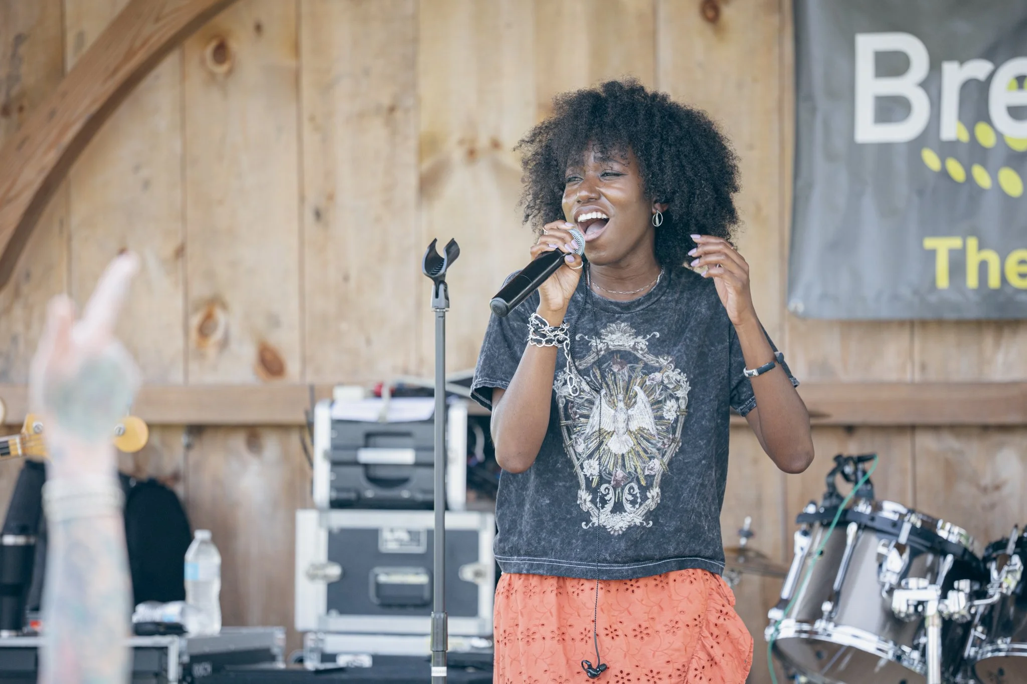 A woman singing into a microphone in a rustic wooden room, with musical equipment and a banner in the background.
