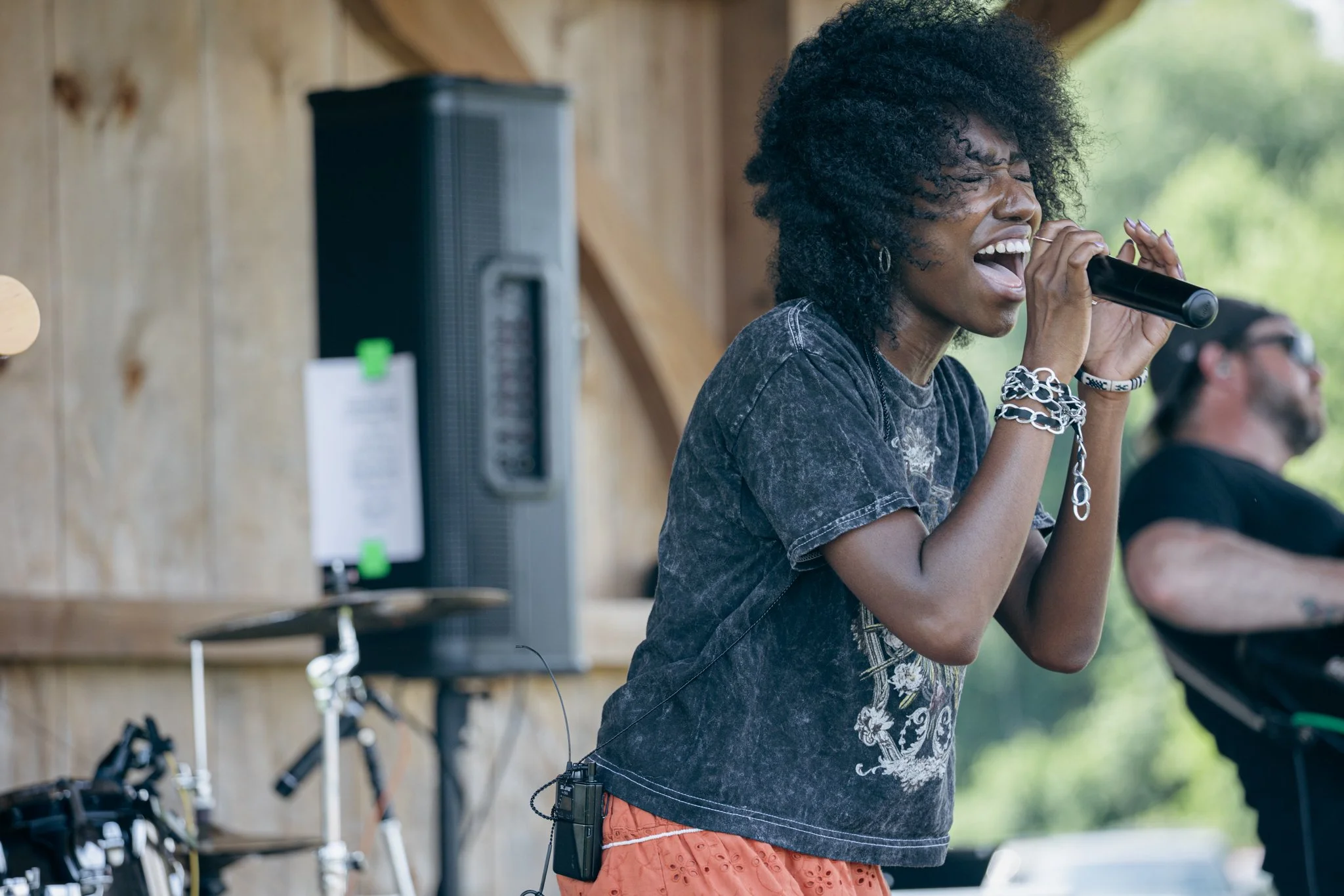 A woman singing into a microphone with a joyful expression, wearing a black graphic t-shirt and orange shorts, with a wood-paneled background and other people and bicycles visible behind her.