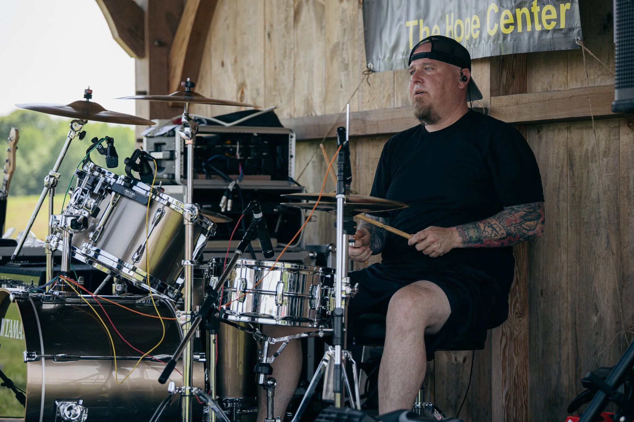 A man with tattoos, wearing a black t-shirt, shorts, and a black cap backwards, sitting outdoors at a drum set, with a wooden wall and a banner that says "The Hope Center" in the background.
