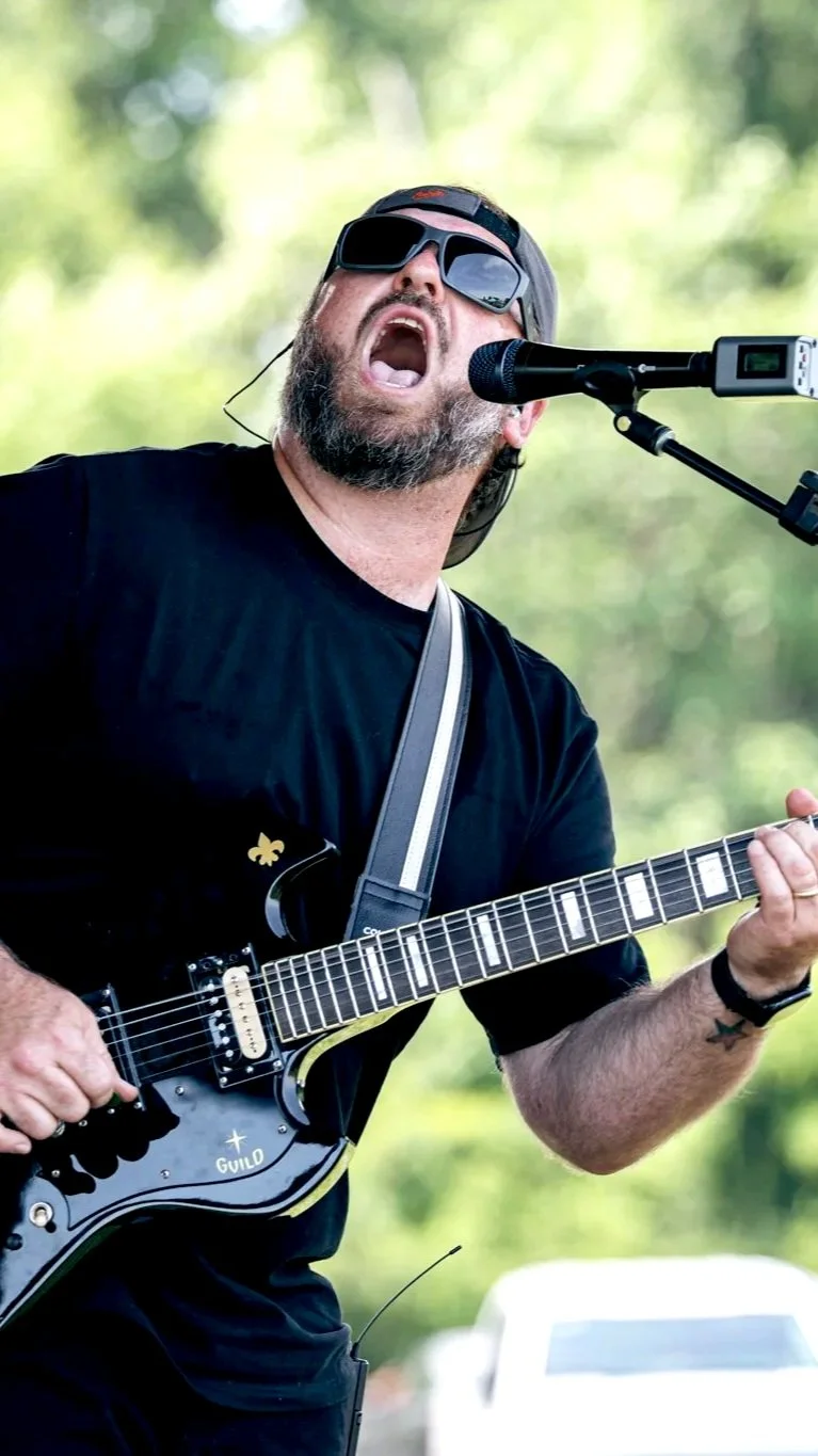 A man singing into a microphone while playing an electric guitar outdoors, wearing sunglasses and a black t-shirt, with a musical earpiece in his ear and a background of green trees.