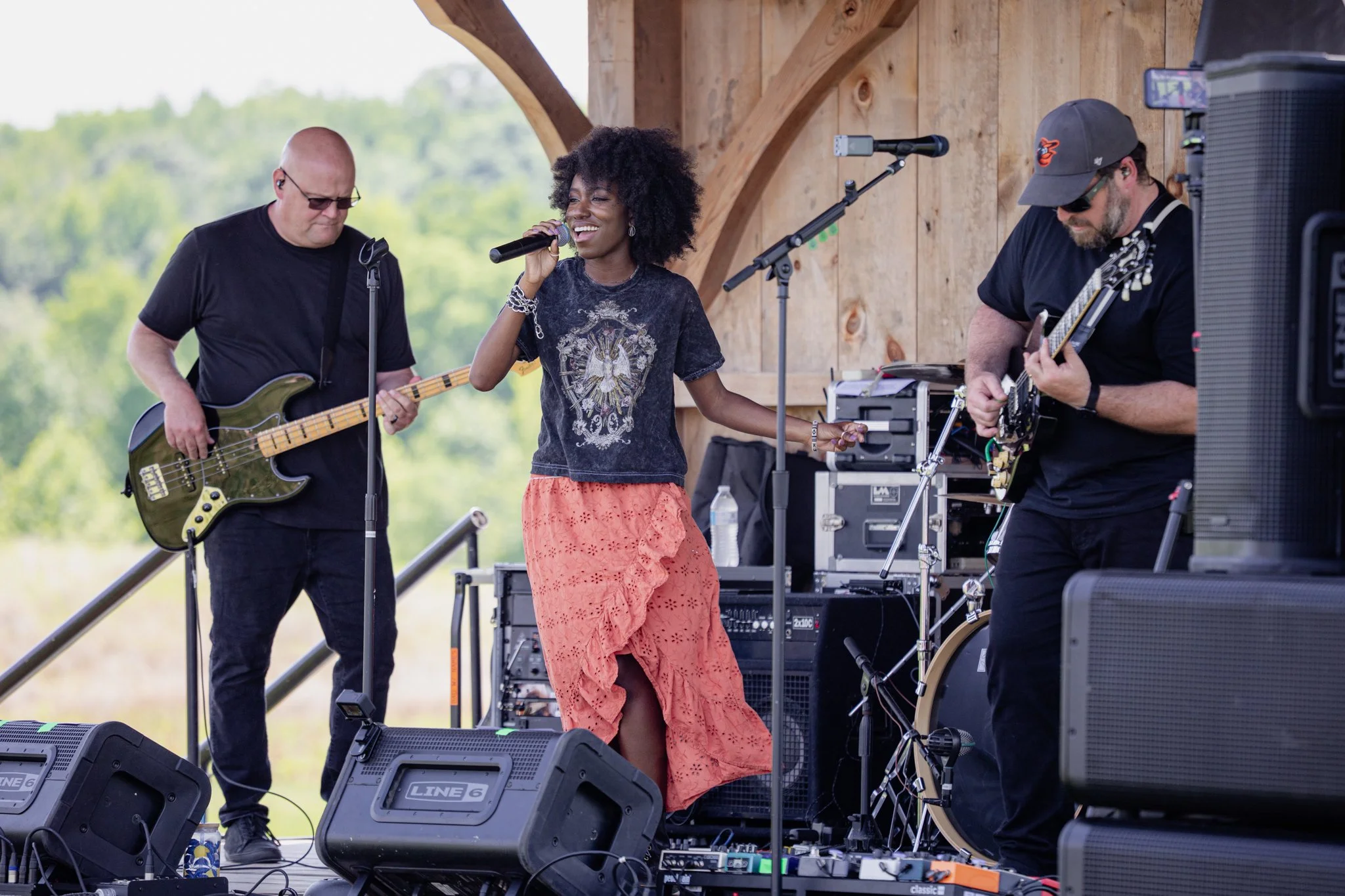 A woman singing into a microphone while two men play guitars beside her on an outdoor stage with a wooden backdrop.