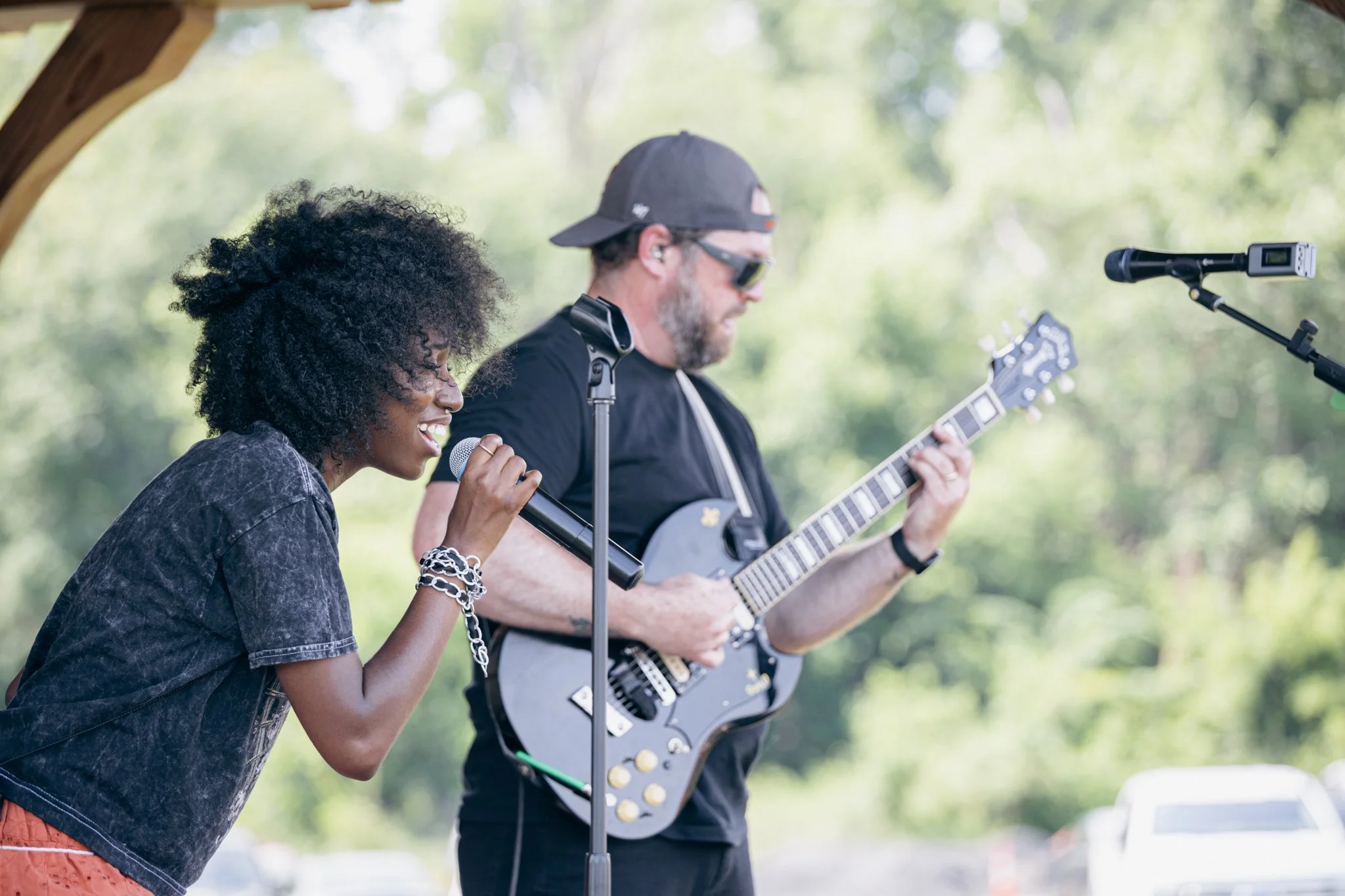 A woman singing into a microphone and a man playing an electric guitar outdoors with blurred green trees in the background.