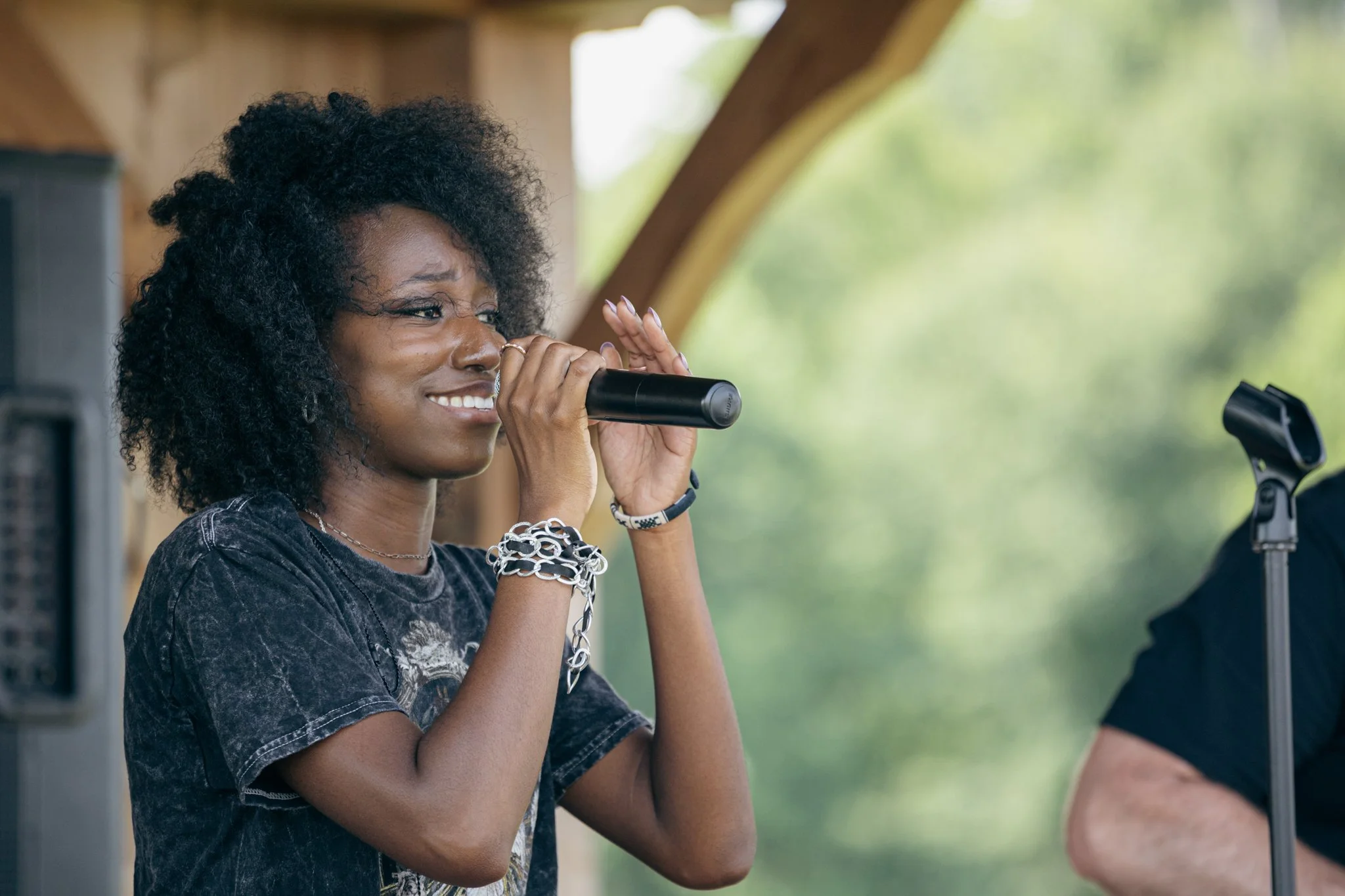 Young woman singing into a microphone outdoors with greenery in the background.