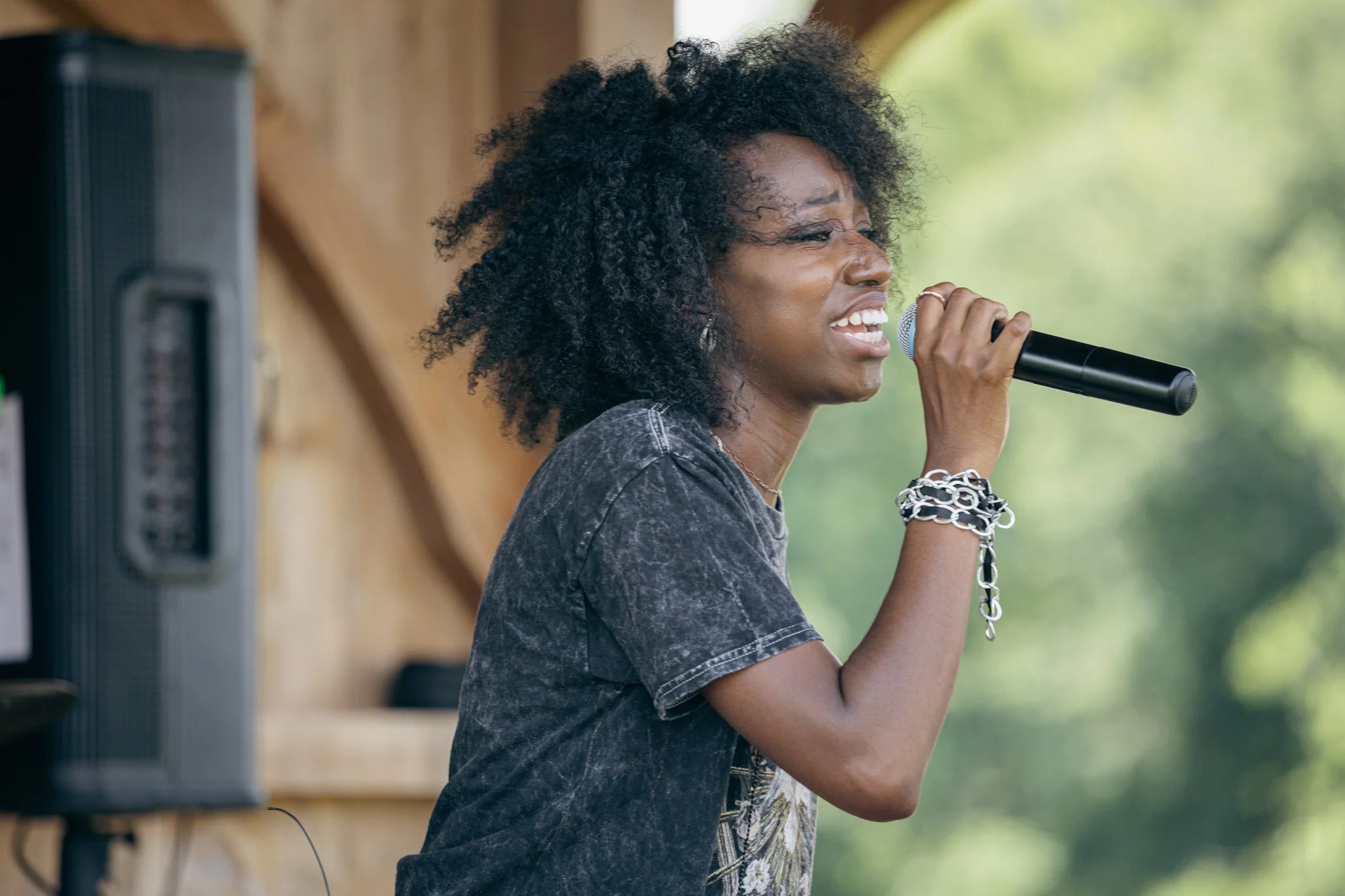 A woman singing into a microphone at an outdoor event with trees and a wooden structure in the background.