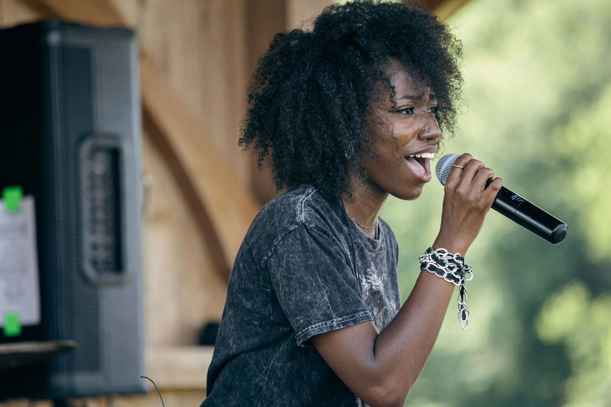 A woman with curly black hair singing into a microphone outdoors, wearing multiple silver bracelets on her wrist.