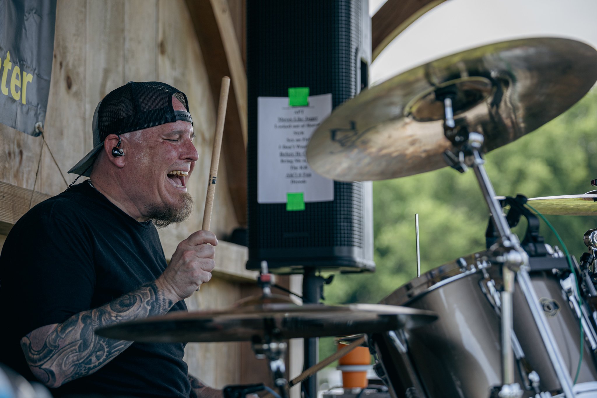 A man with tattoos, wearing a black baseball cap and black shirt, is playing drums outdoors, smiling passionately with cymbals and drum equipment around him.